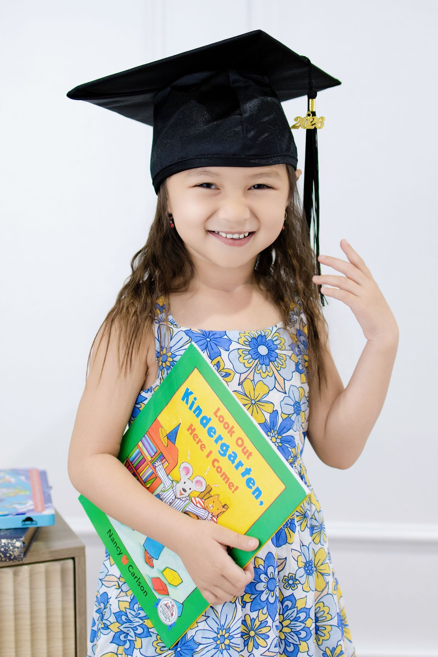 Young girl in graduation cap holding a Kindergarten book, smiling proudly in a floral dress.