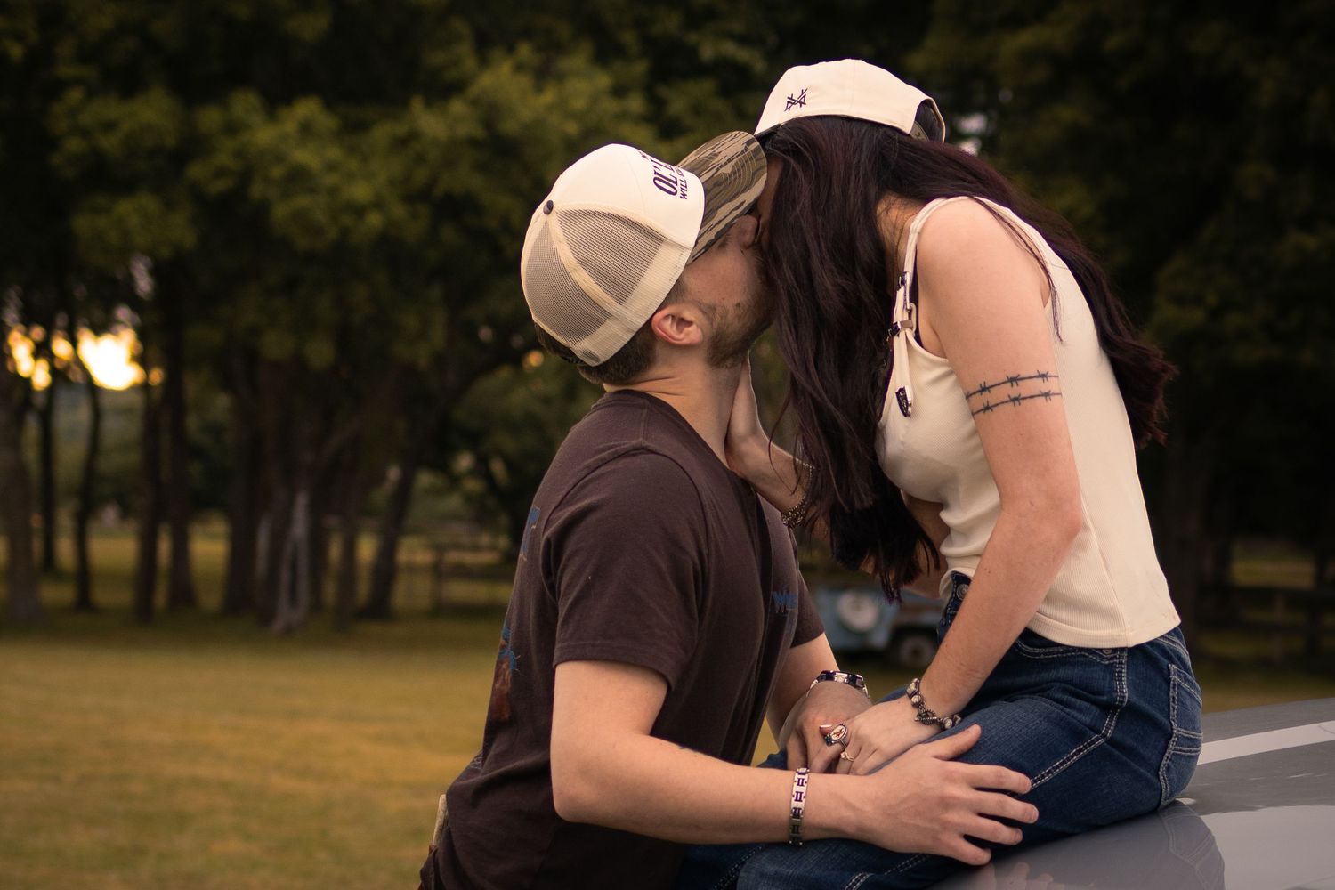 A young couple sharing a romantic kiss outdoors, both wearing white caps, with lush green trees in the background.