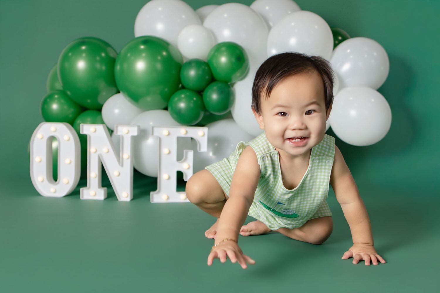 Baby smiling and crawling in front of green and white balloon garland with illuminated ONE letters for first birthday.