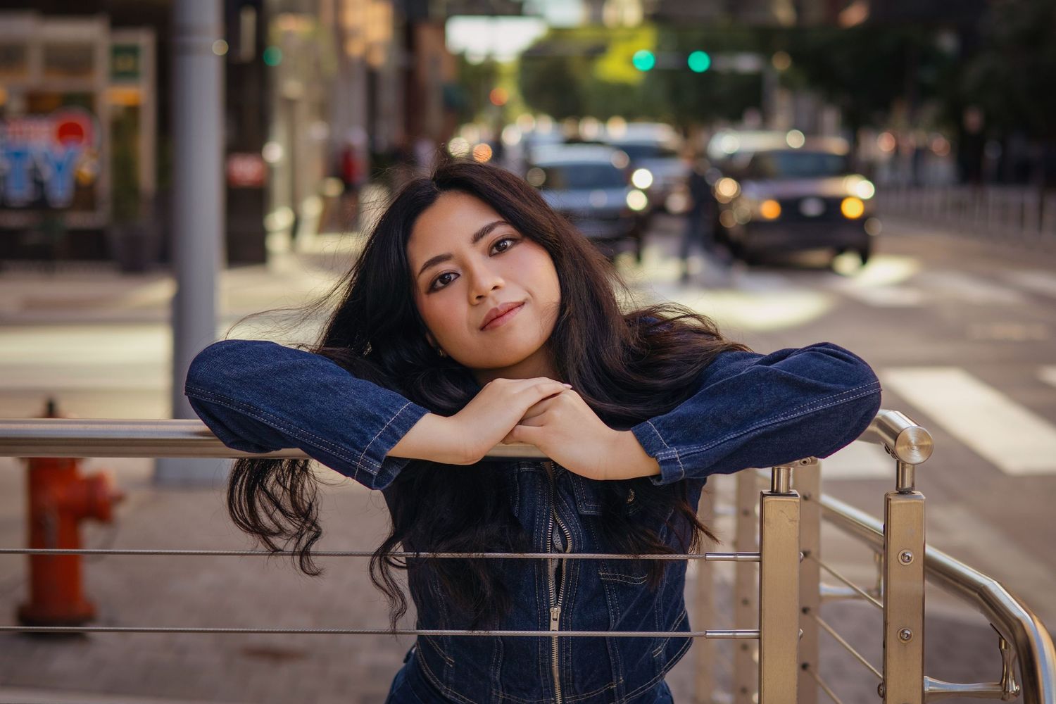 Young woman in denim jacket leaning on railing on city street.