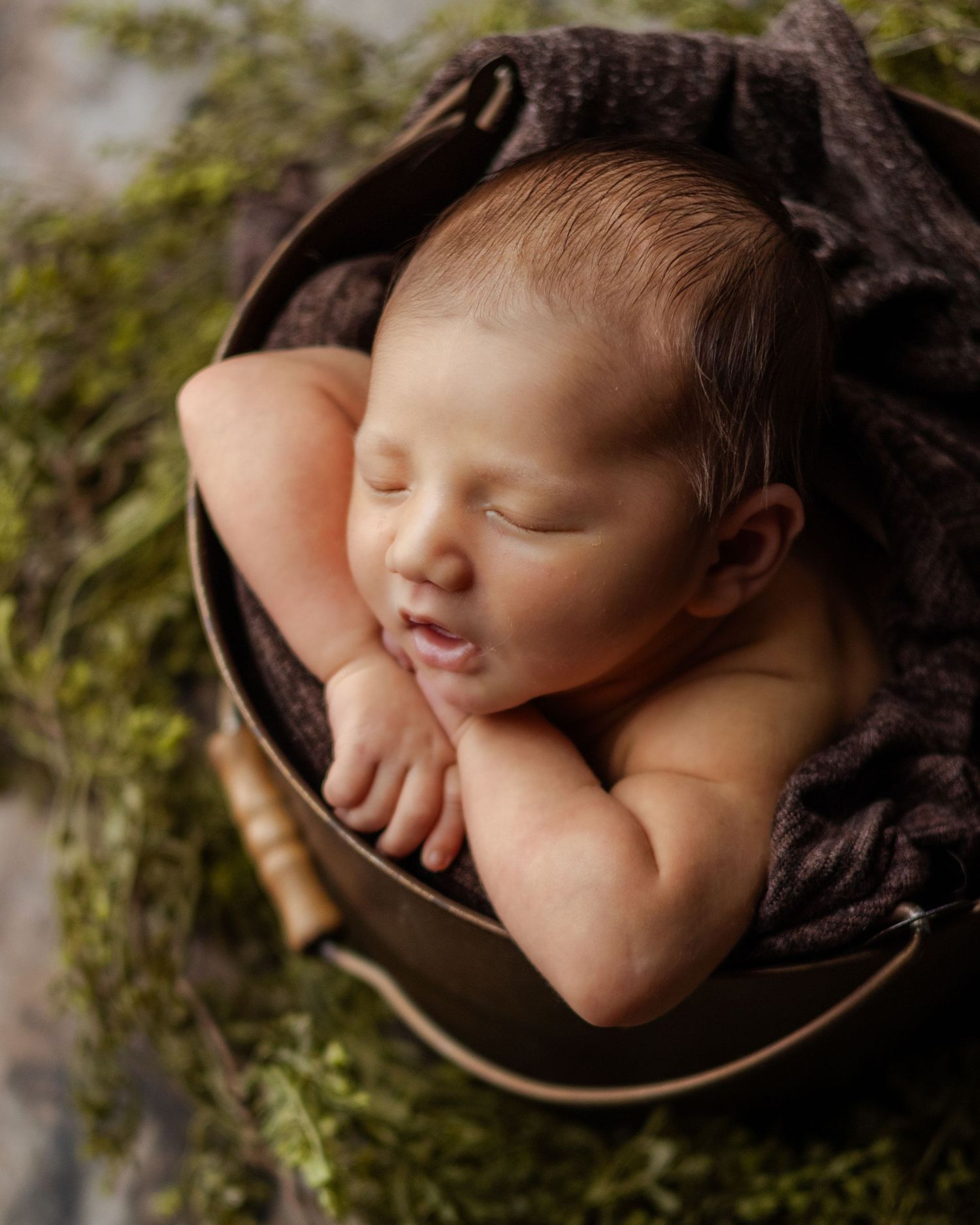 Sleeping newborn baby curled in a rustic bucket with dark fabric and green moss, peaceful newborn photography.