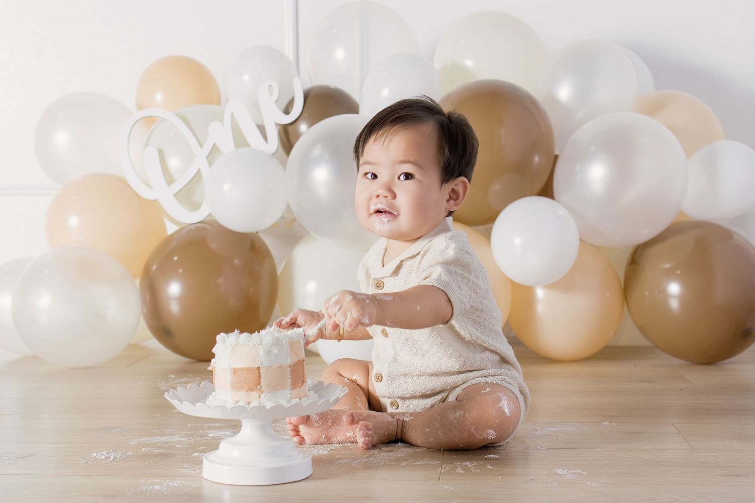 Baby's first birthday cake smash with gold and white balloons, neutral beige decor, and a smiling infant in a cream romper.