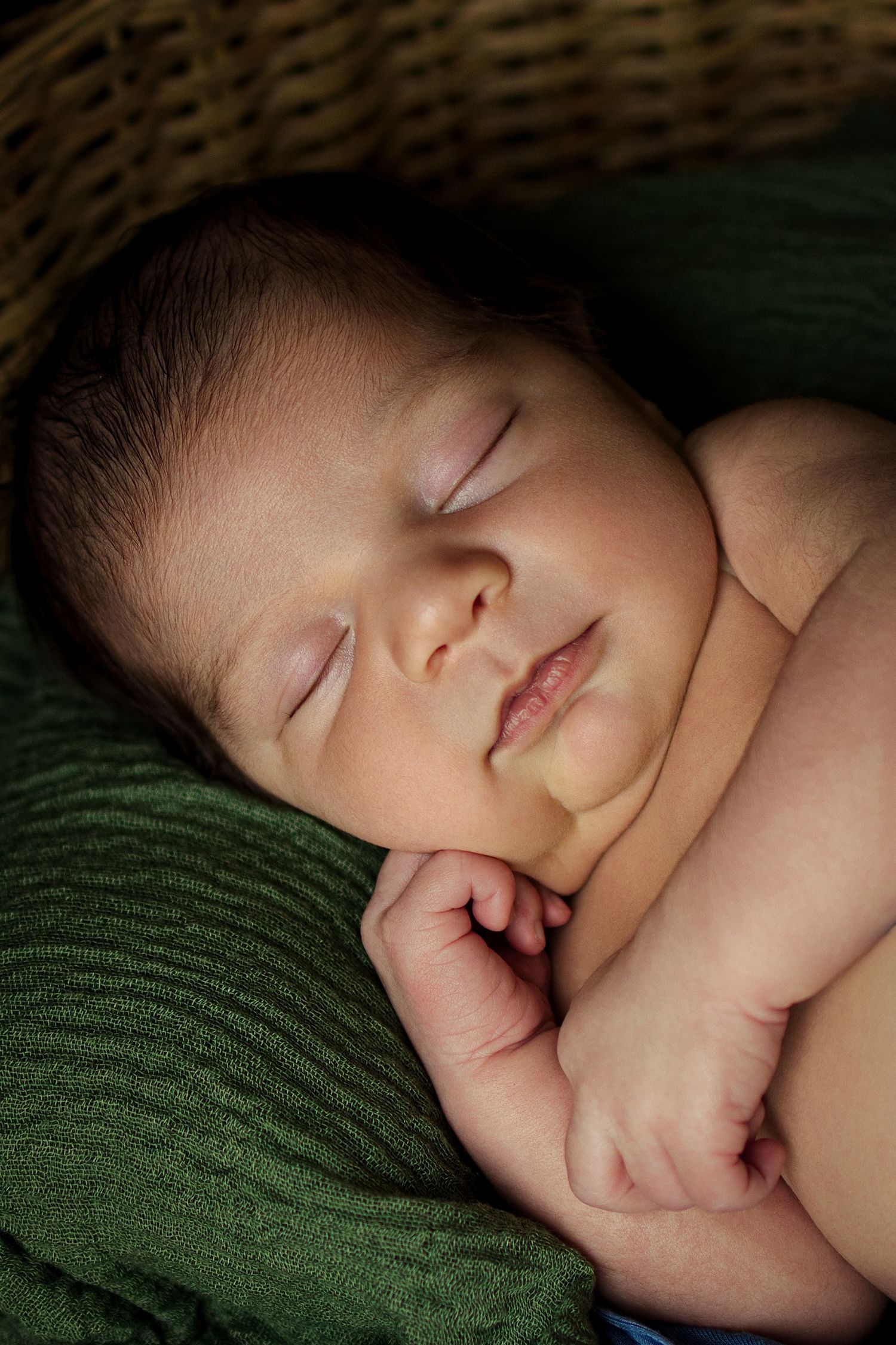 Peaceful newborn baby sleeping curled up on a green blanket inside a wicker basket.
