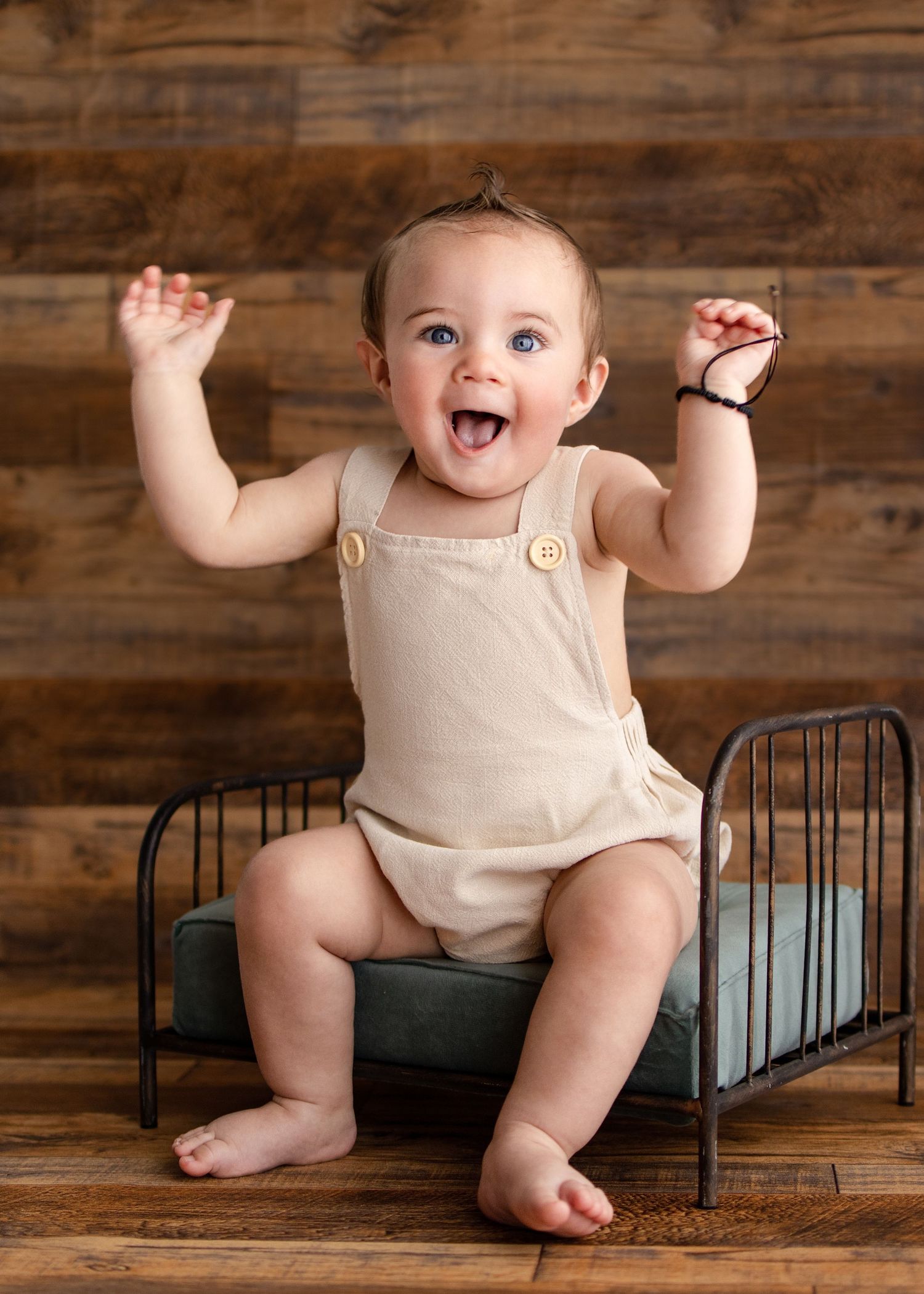Happy baby in beige romper sitting on mini bed with arms raised, laughing joyfully.