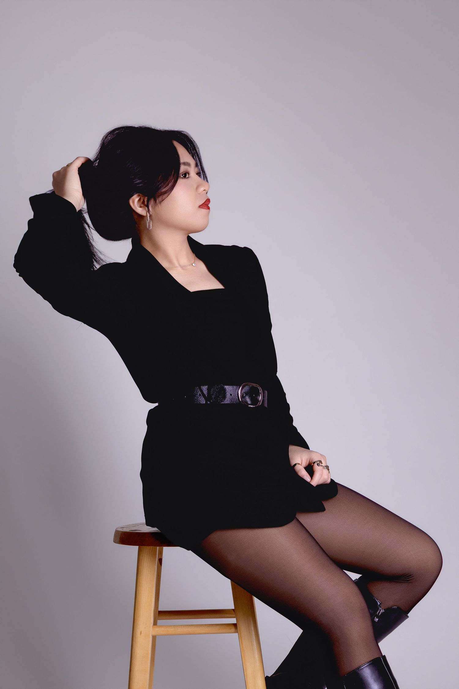 Young woman in black belted dress seated on stool, hand in hair, red lips, studio fashion portrait.