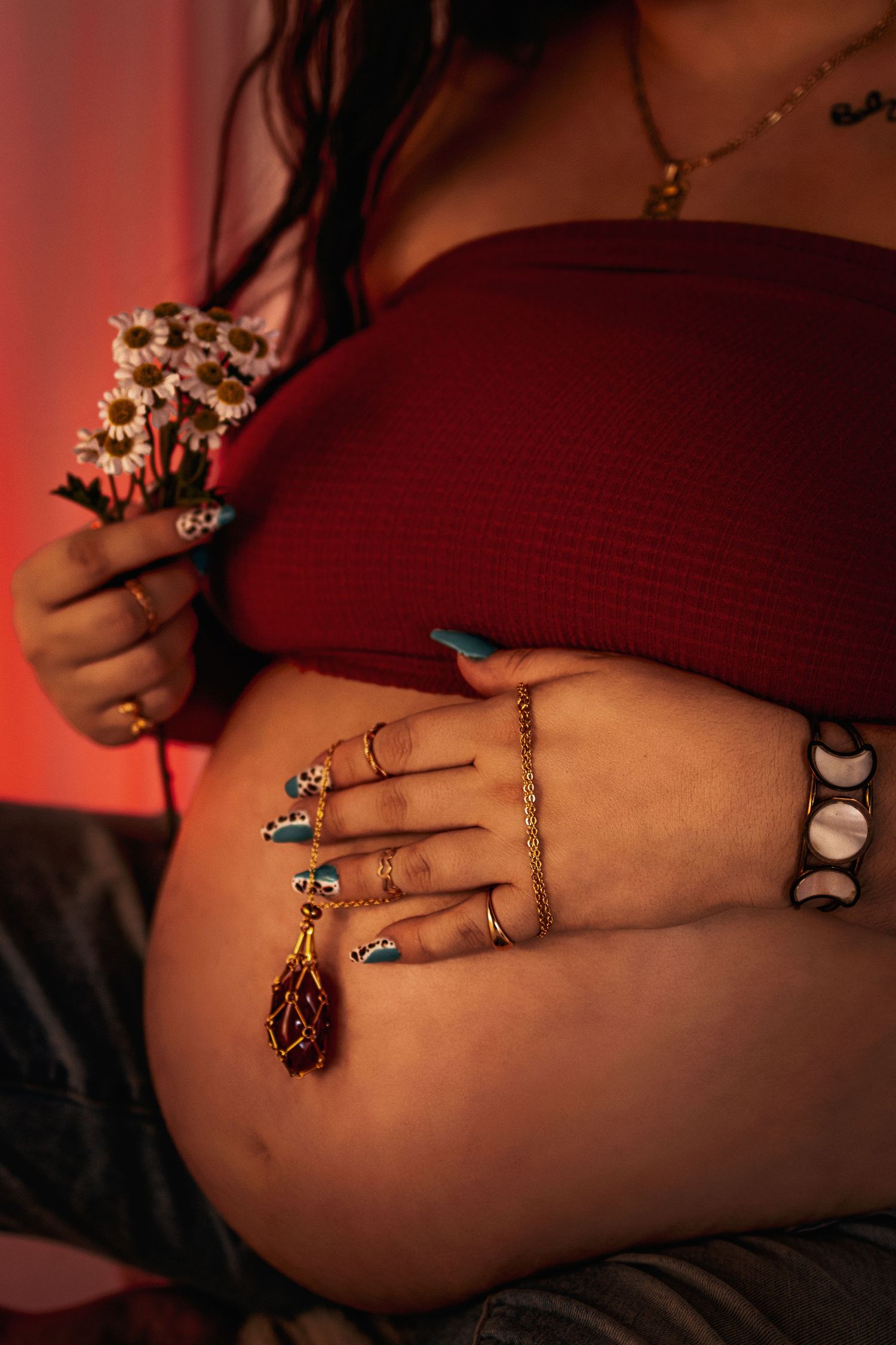 Pregnant woman in red crop top with rings and gold jewelry resting hand on bare belly, holding small white flowers.
