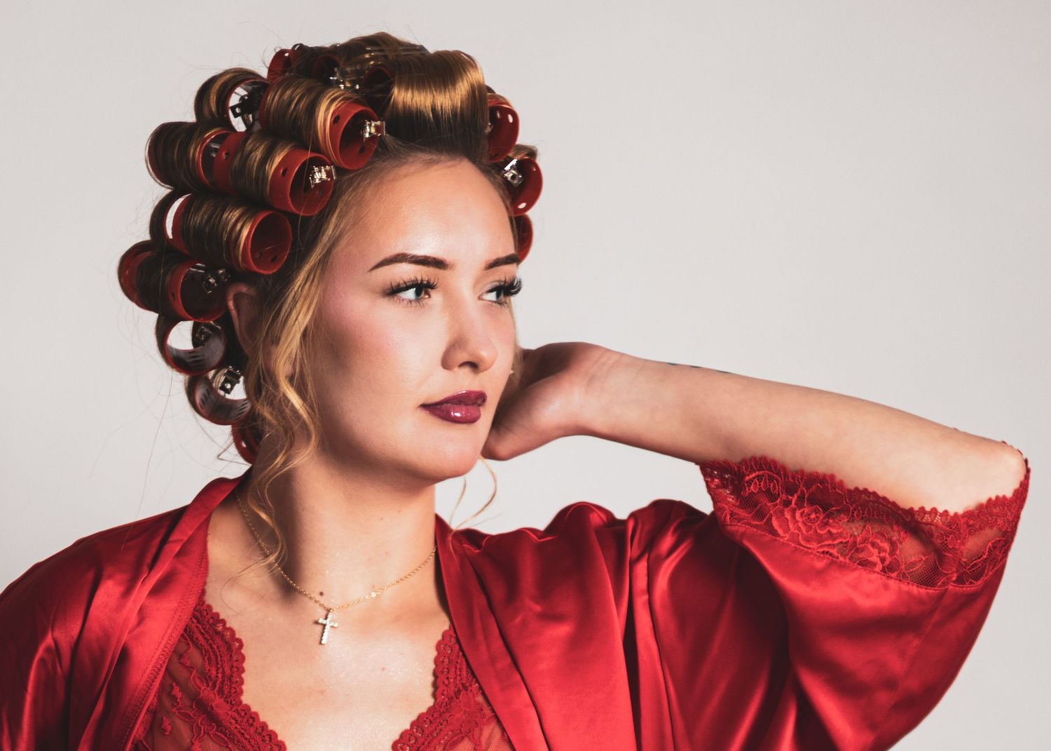 Young woman in red satin robe with red hair rollers poses elegantly against a light gray background.