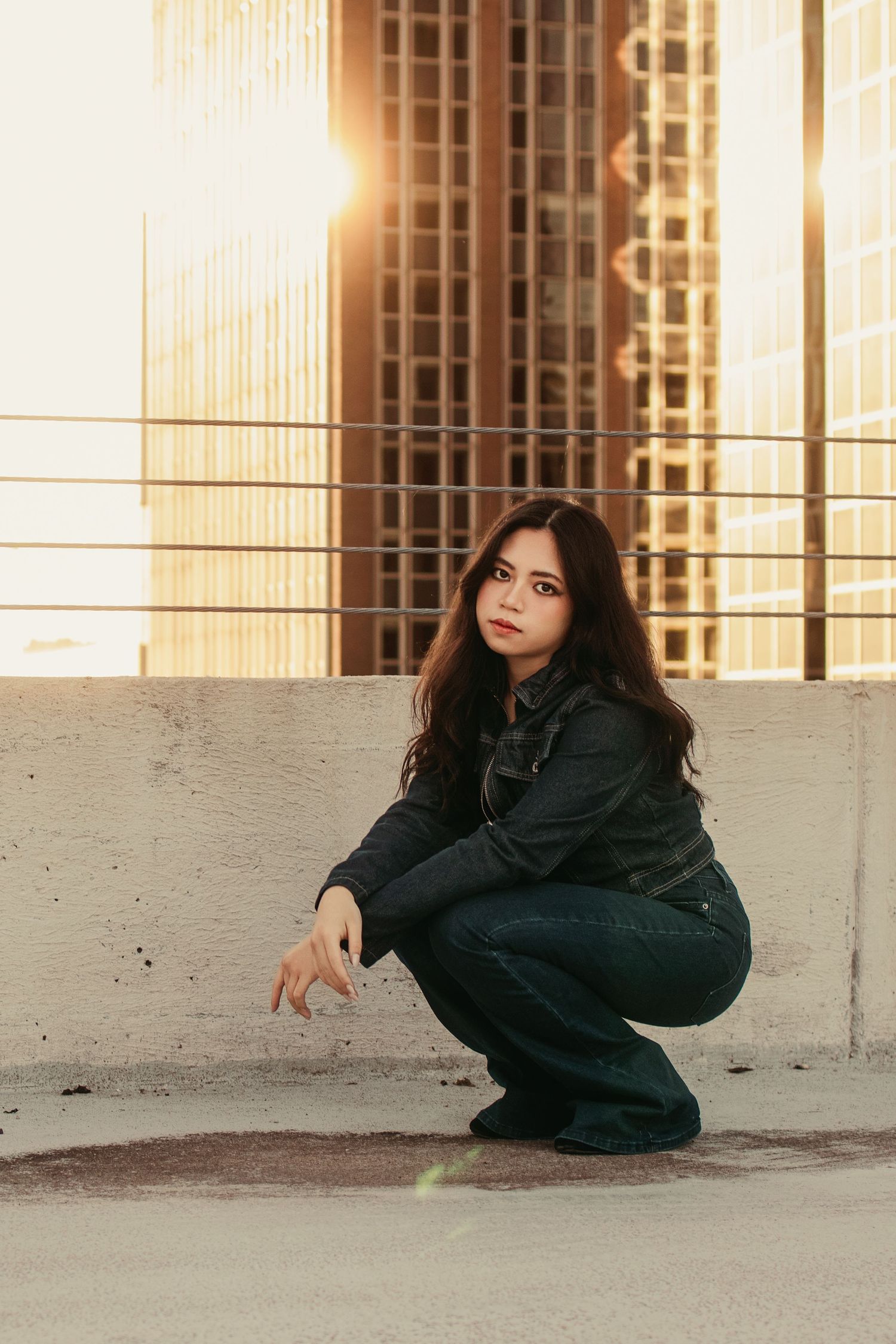 Young woman in denim crouches on rooftop with golden sunset reflecting off city skyscrapers.