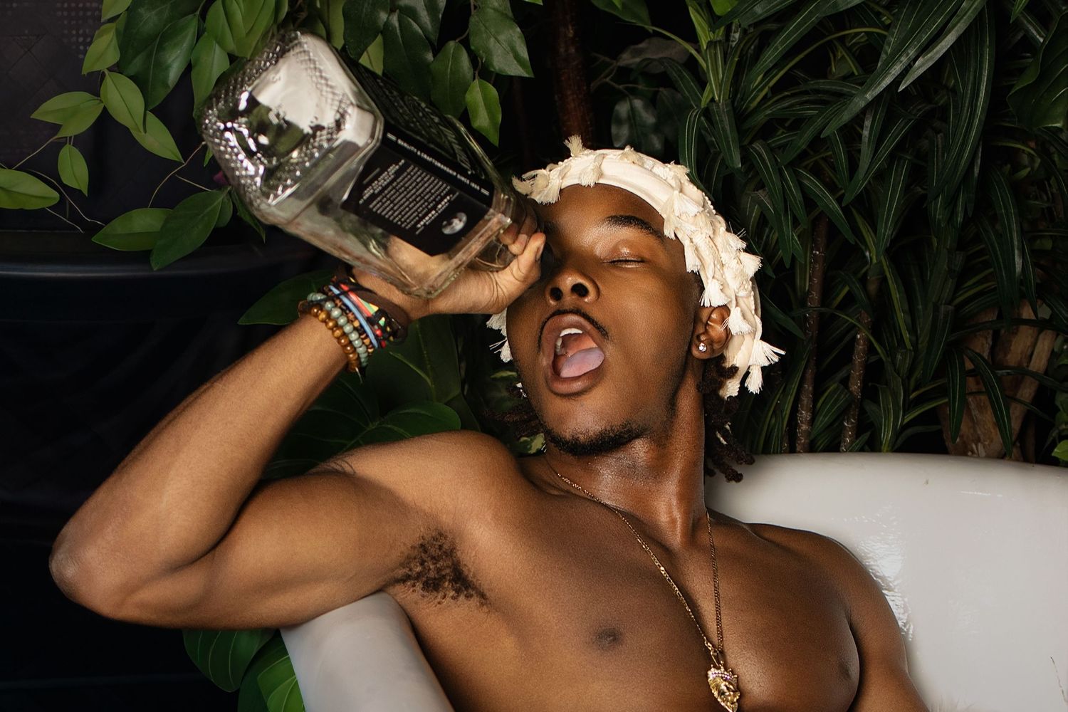 Shirtless young man drinking from a bottle while relaxing in a white chair surrounded by tropical plants.