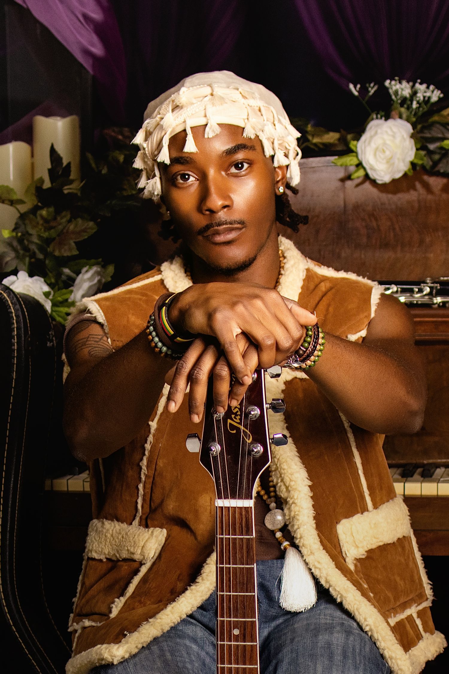 Young Black musician in shearling vest and head wrap rests chin on acoustic guitar neck indoors.