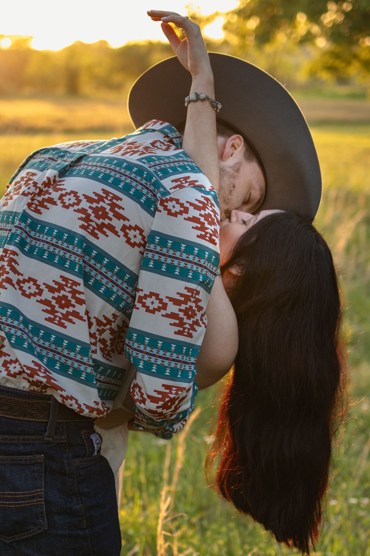 Couple sharing a romantic dip kiss outdoors at golden sunset, man in cowboy hat and Aztec print shirt, woman with long dark hair.