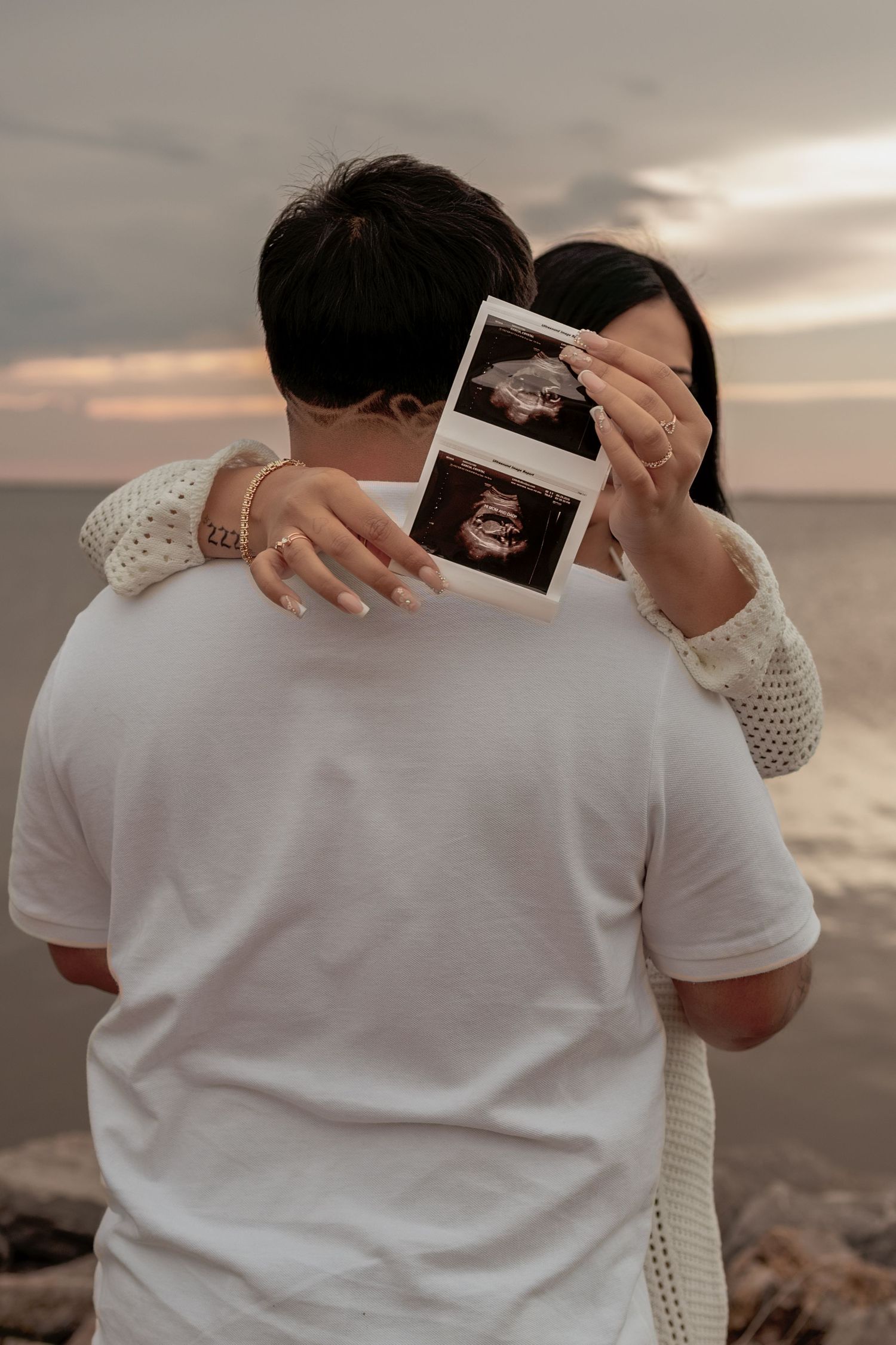 Couple embracing at sunset beach, holding ultrasound photos, announcing pregnancy near rocky shoreline.