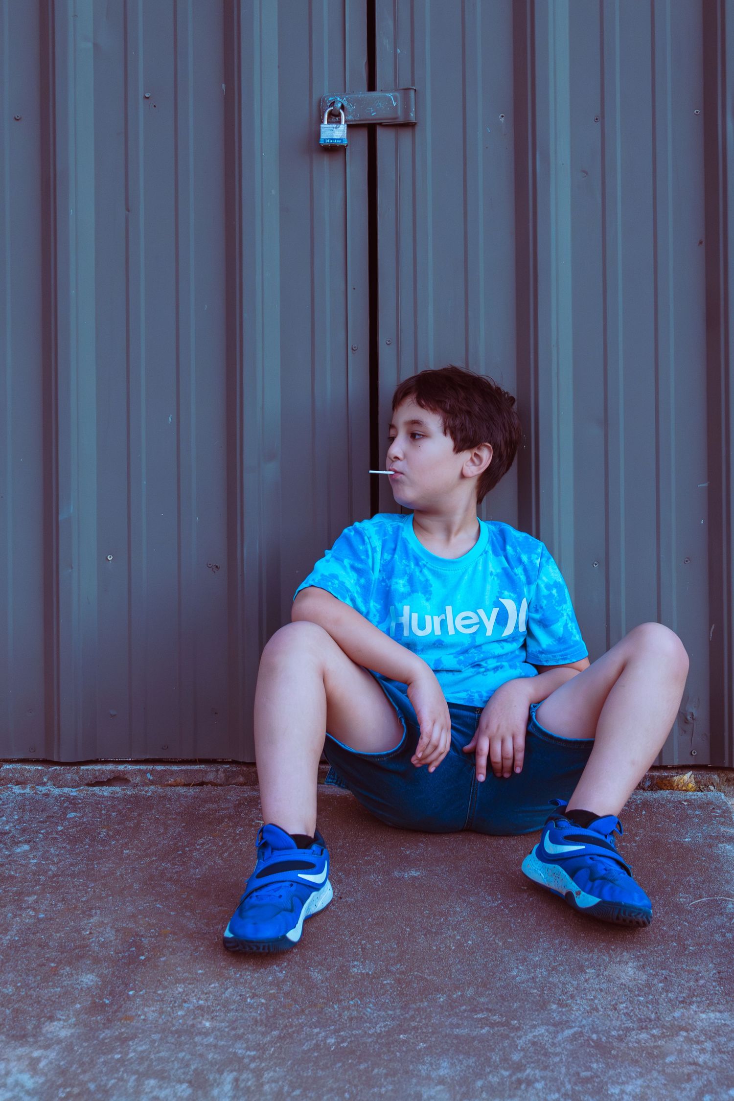 Young boy in blue Hurley shirt and shorts sitting on skateboard against metal wall with padlock.