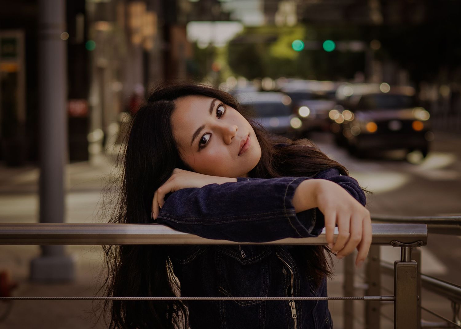 Young woman with long dark hair leans on a railing, gazing upward on a city street at dusk.