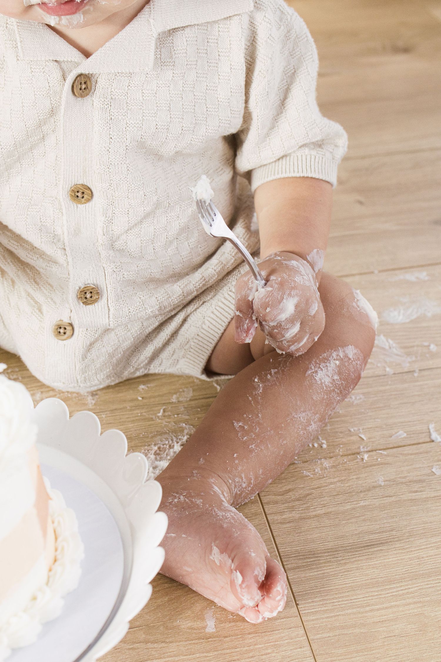 Baby in beige knit outfit sits on wood floor holding fork with cake smash mess on legs beside white birthday cake.