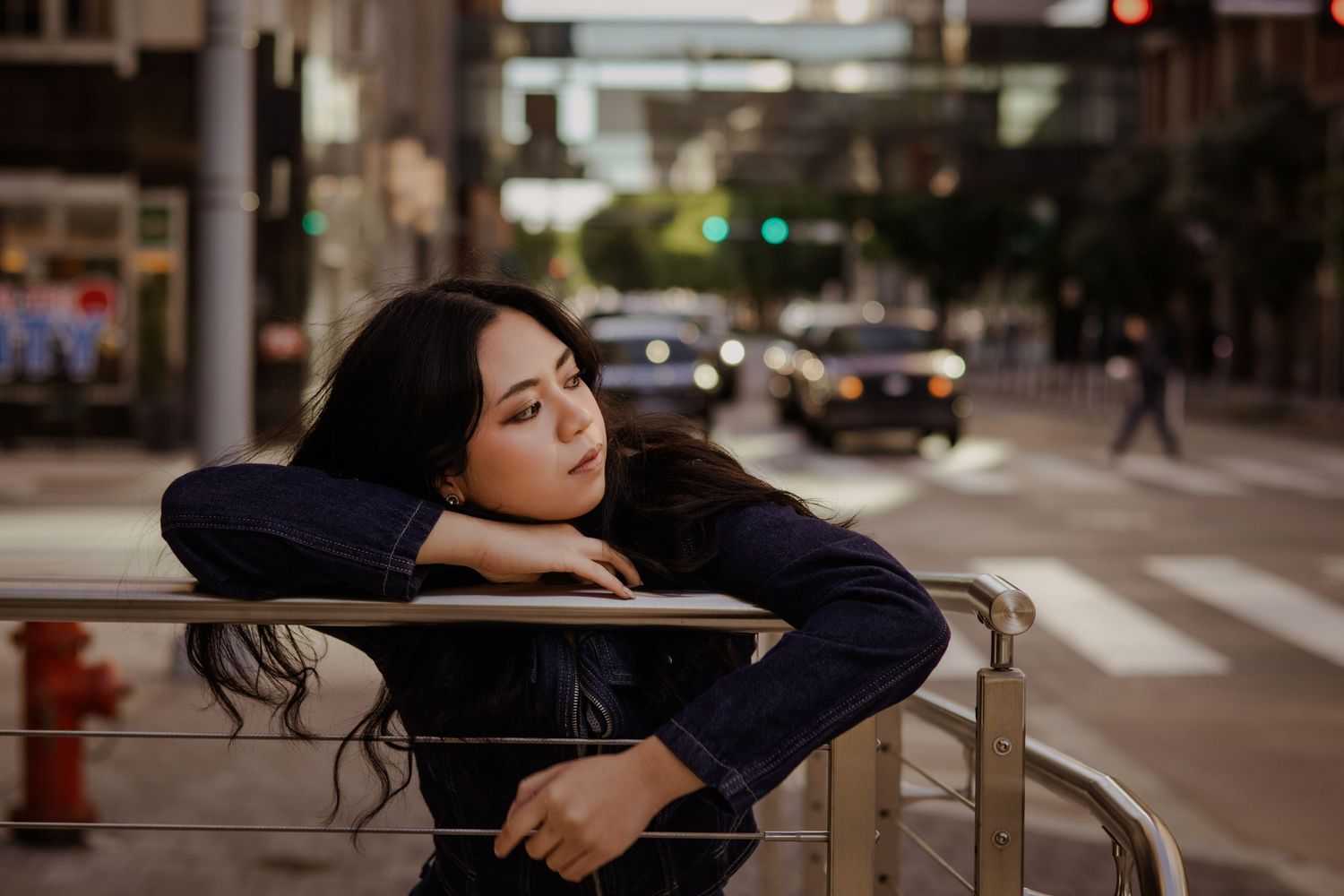 A young woman with dark hair leans on a metal railing on a city street, looking contemplative near a crosswalk.