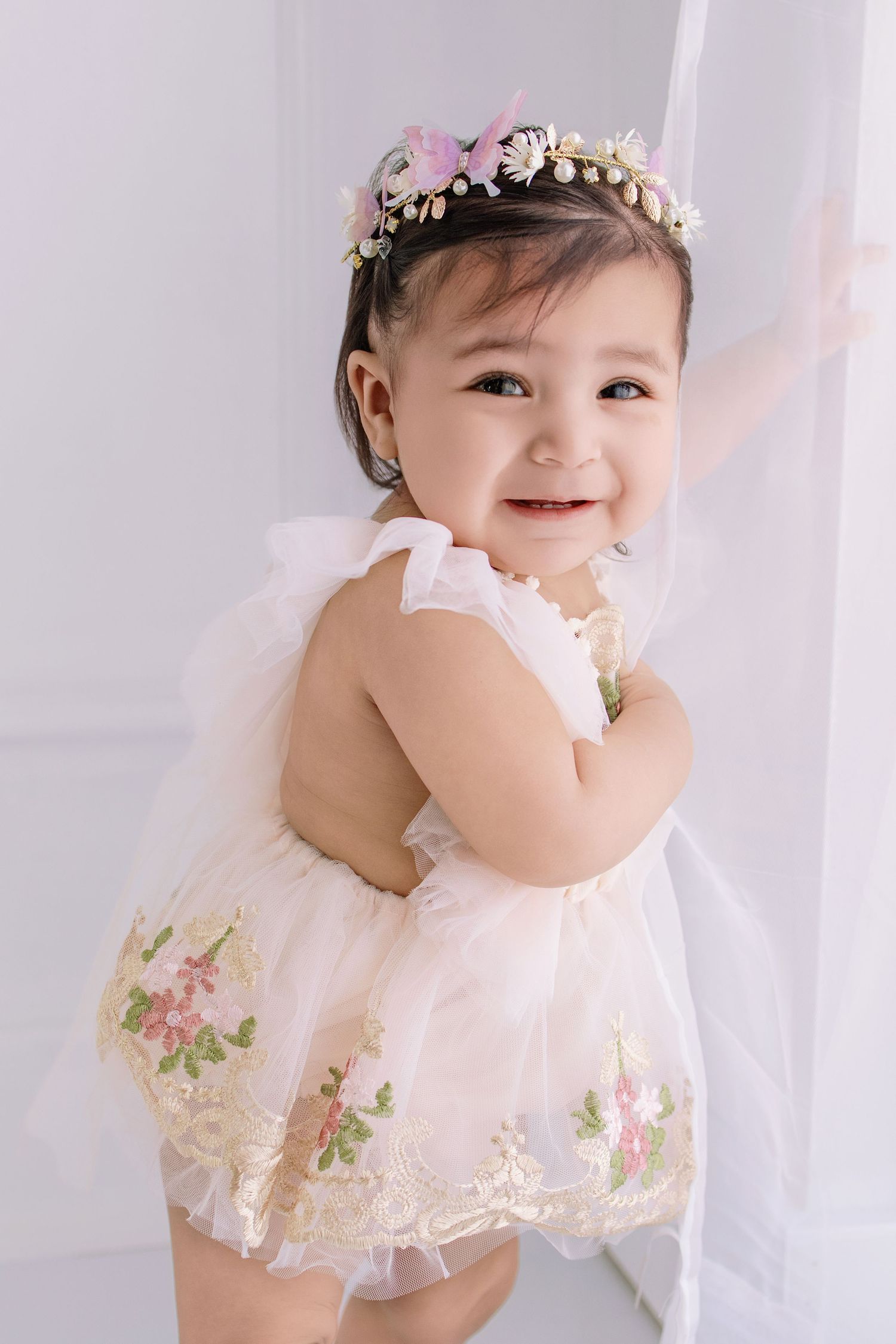 Smiling baby girl in white floral embroidered dress and colorful flower crown standing by curtain.