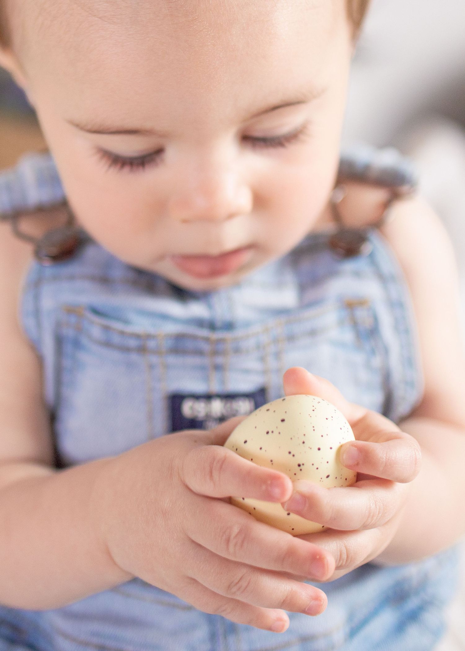 Cute baby in denim overalls carefully holding a small speckled Easter egg in tiny hands.