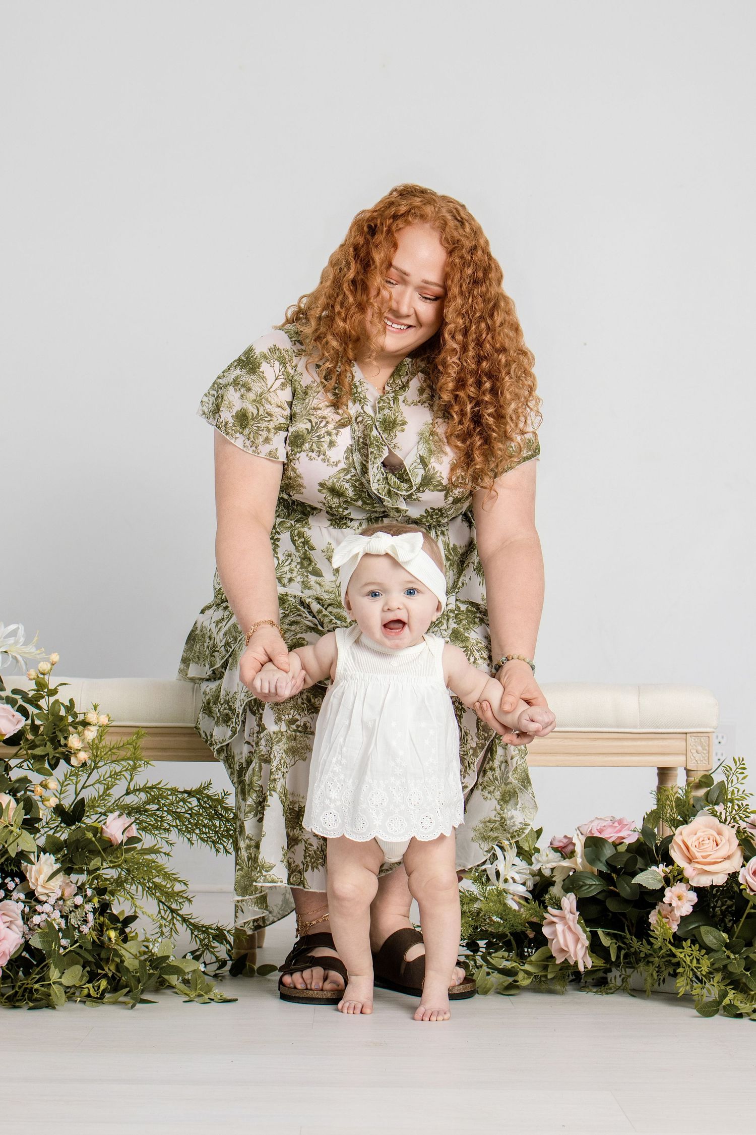A smiling redhead woman in floral dress helps a baby girl in white dress stand among pink flowers.