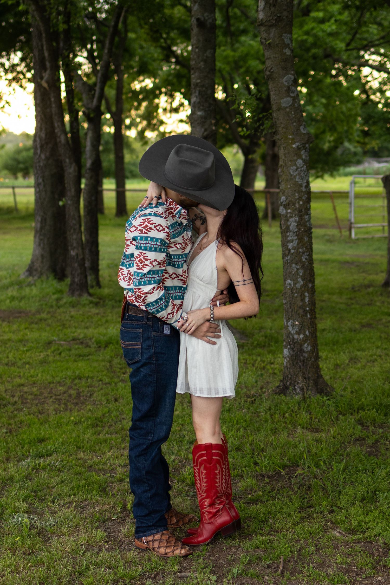 Couple sharing a kiss under trees, woman in white dress and red cowboy boots, man in aztec print shirt.