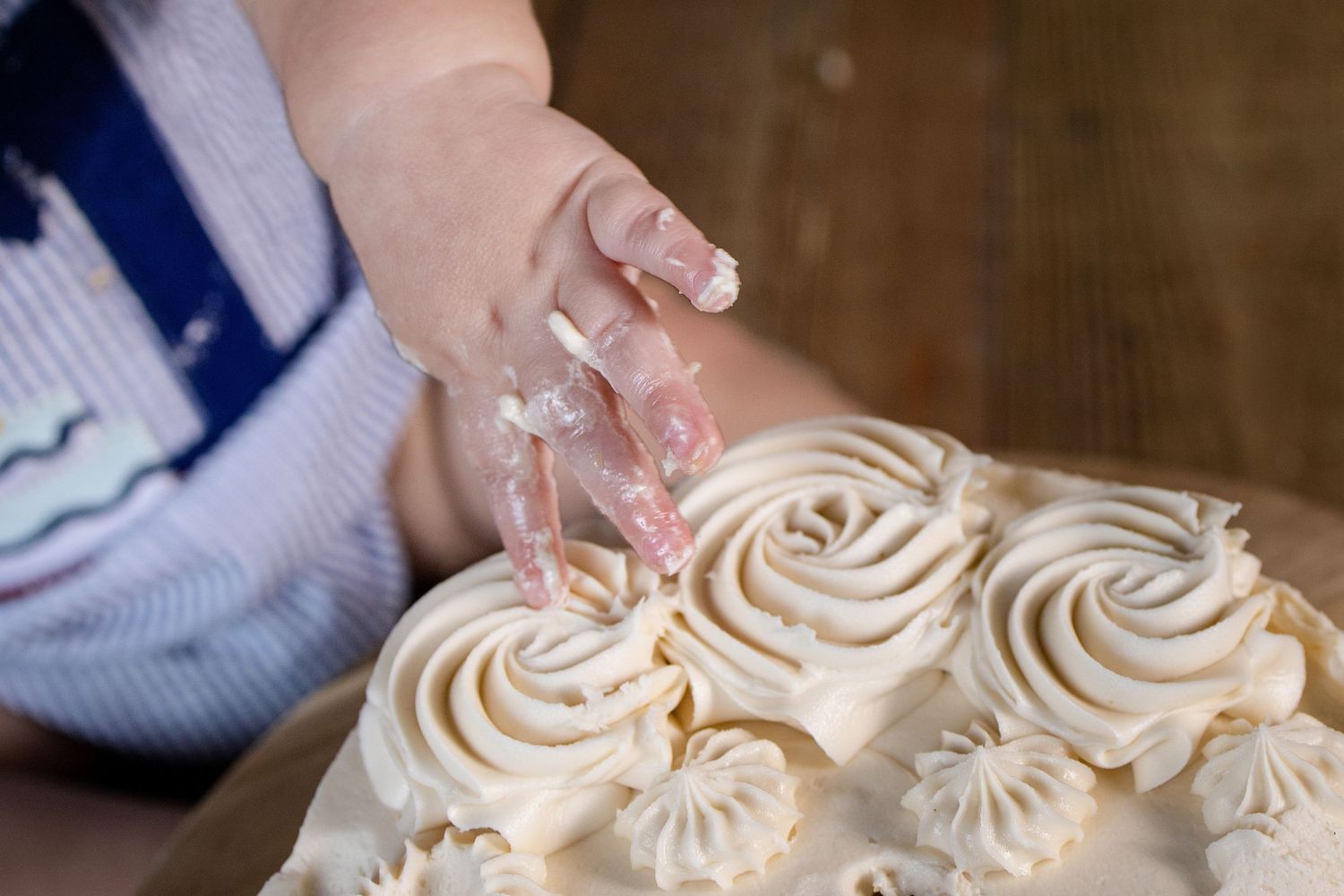 A child's hand reaching into a cake decorated with swirled white meringue rosettes on a wooden surface.