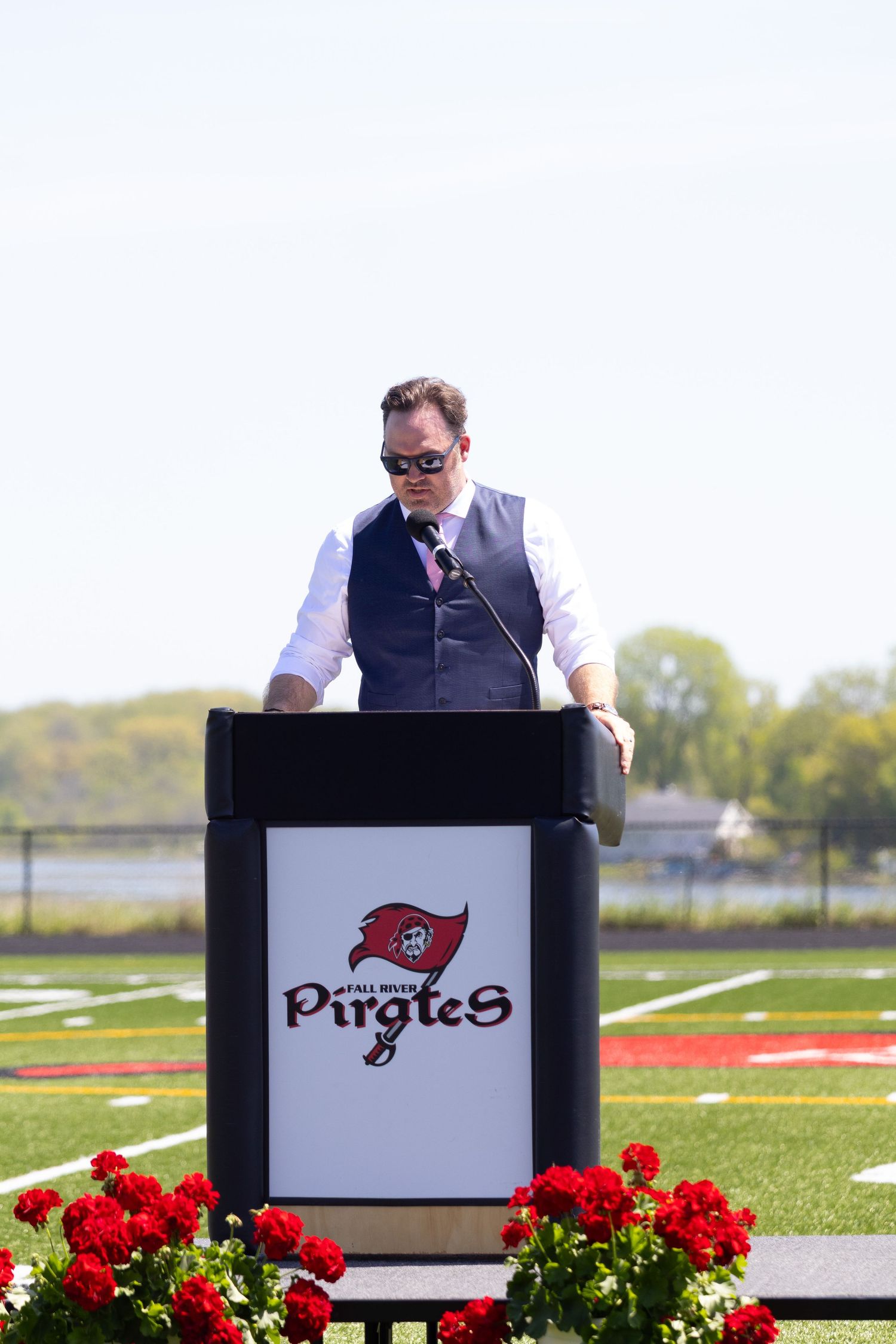 Man speaking at a Pirates podium on a football field, wearing a vest and tie, with red flowers in the foreground.