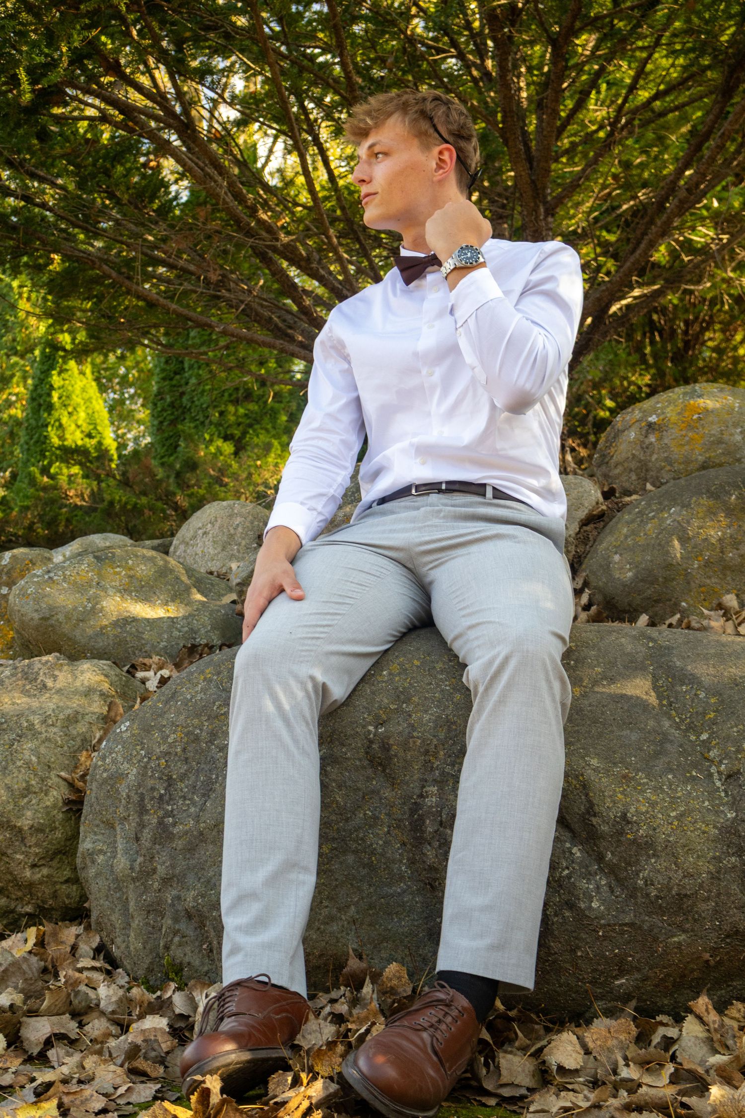Young man in white shirt, bow tie, and gray trousers sitting on rocks in a garden setting.