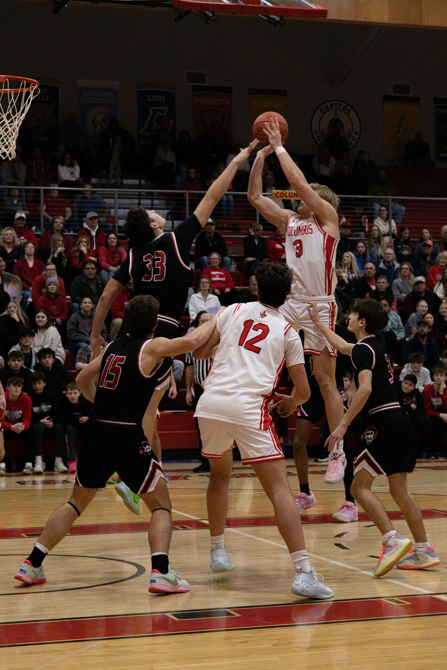 Basketball players compete for a rebound during an intense indoor game between teams in red and black uniforms.