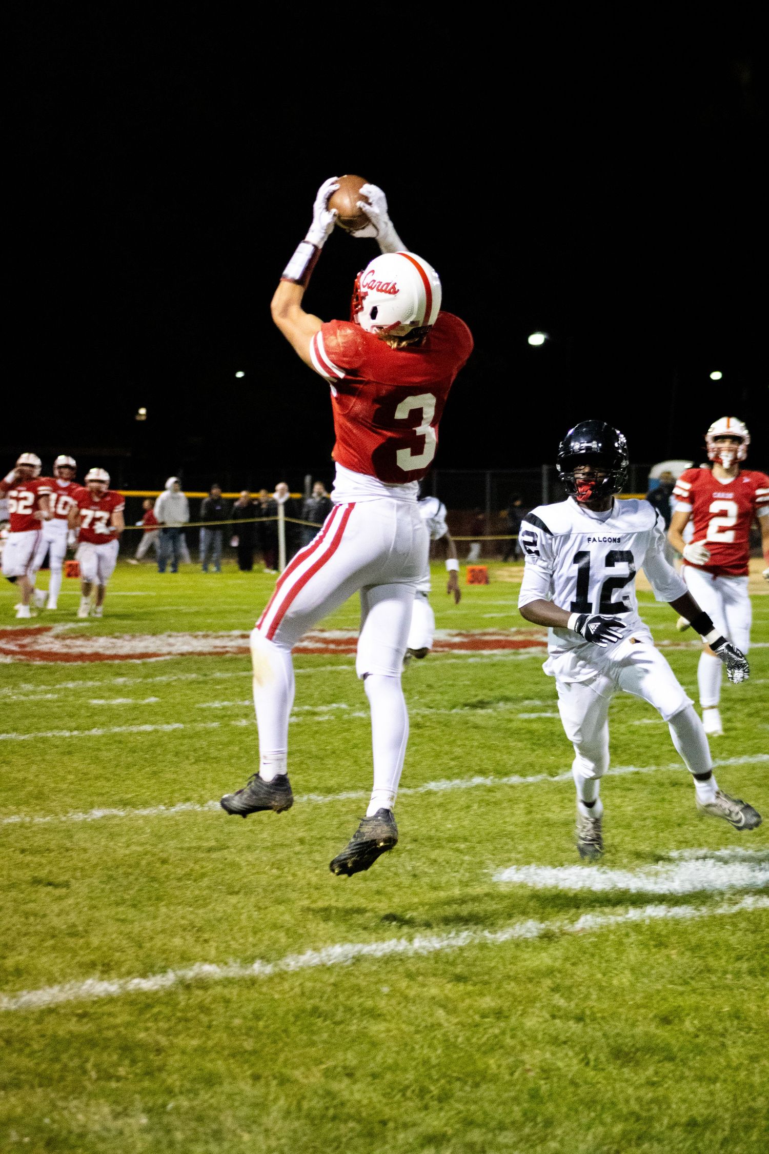 High school football player in red jersey makes leaping catch during night game as defender in white jersey pursues.