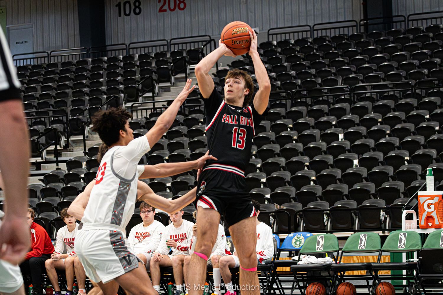 Basketball players compete for a rebound during an indoor game with empty stadium seats in the background.