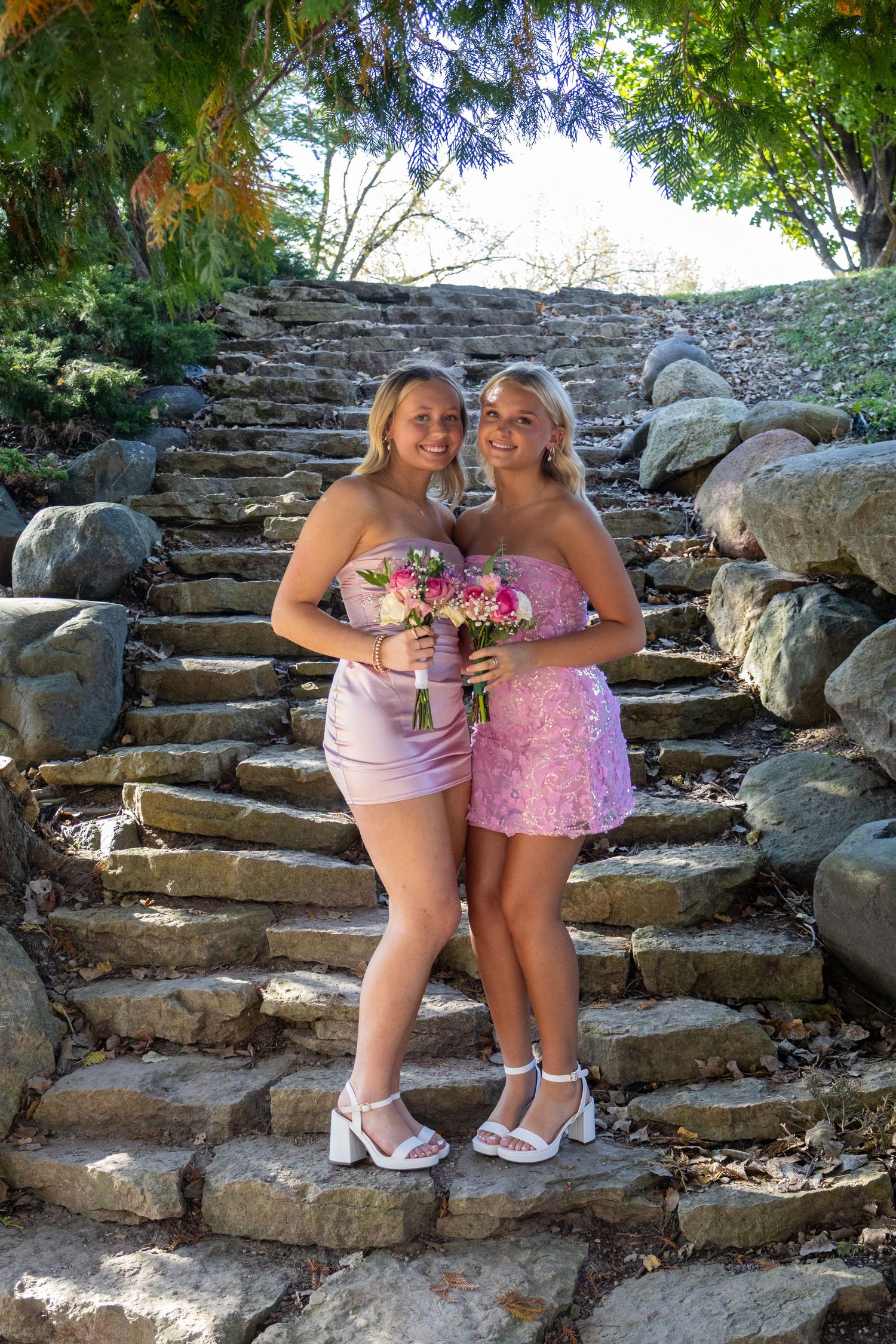 Two blonde teenage girls in pink dresses holding floral bouquets pose on stone steps surrounded by lush greenery.