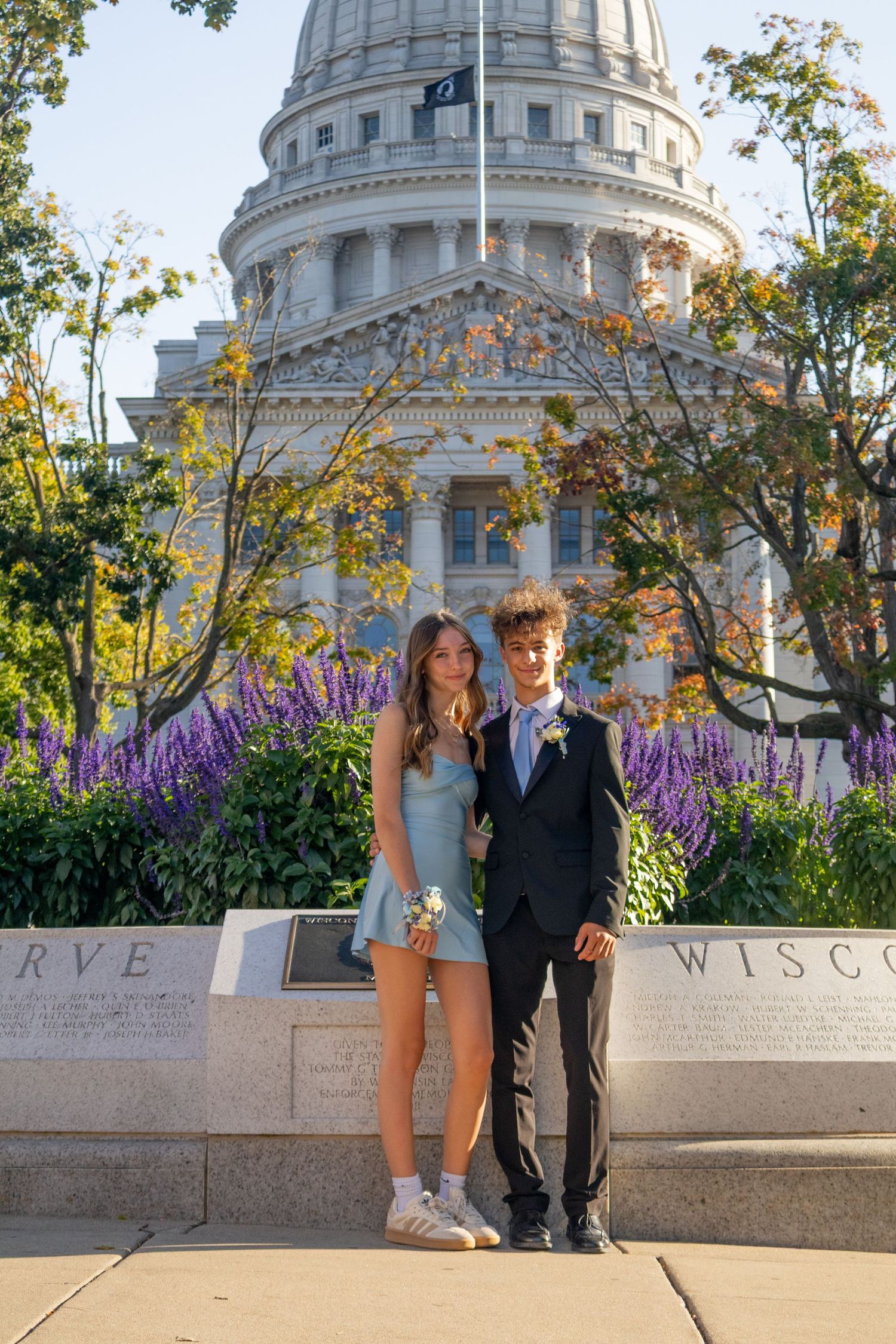 A couple poses in formal attire in front of the Wisconsin State Capitol building with purple flowers in the background.