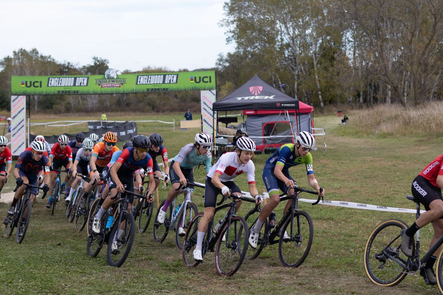 Cyclists compete in a cross-country mountain bike race on a grassy course with banners and spectators in the background.