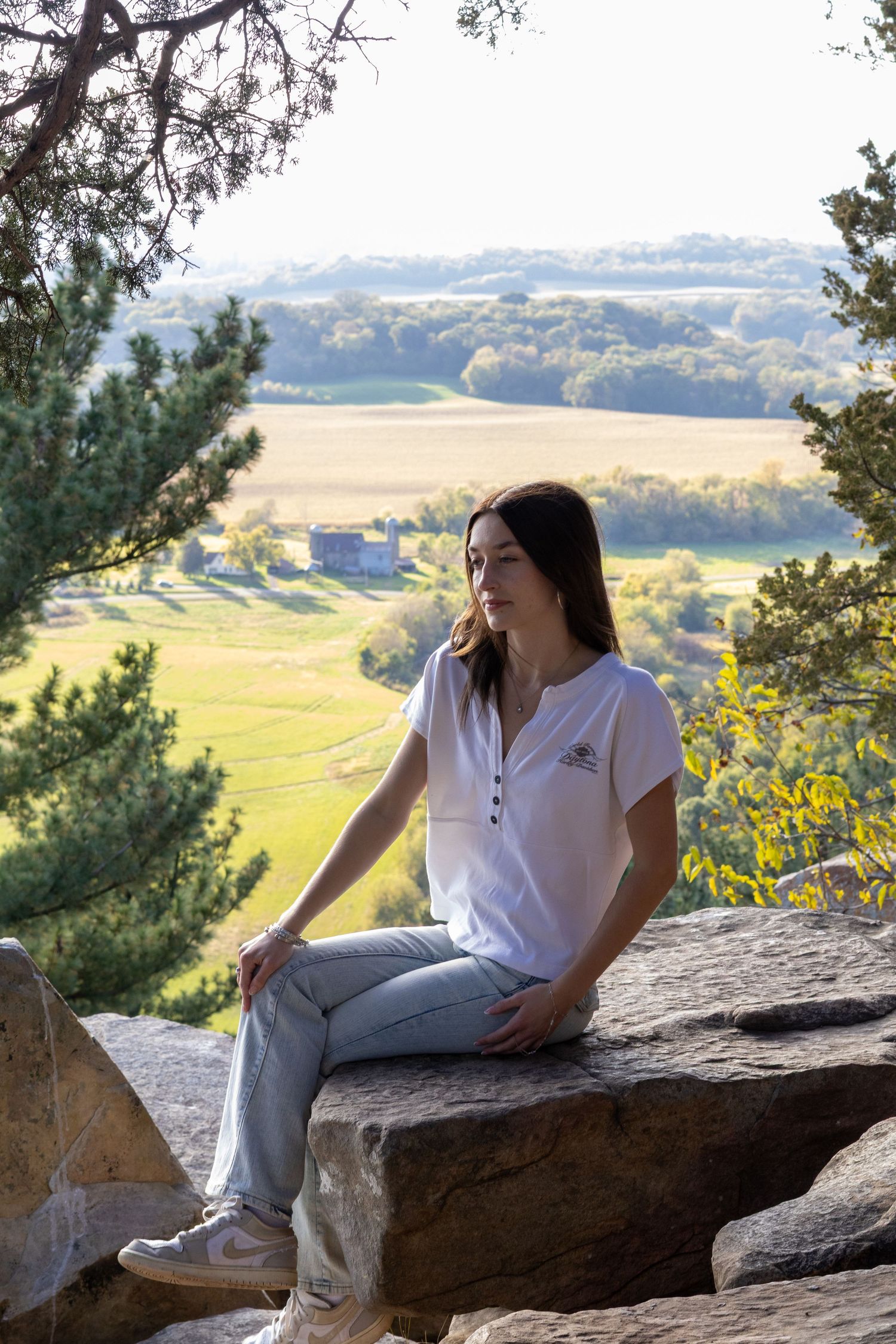 Young woman sitting on rocks at scenic overlook with green fields and rolling hills in background on sunny day.