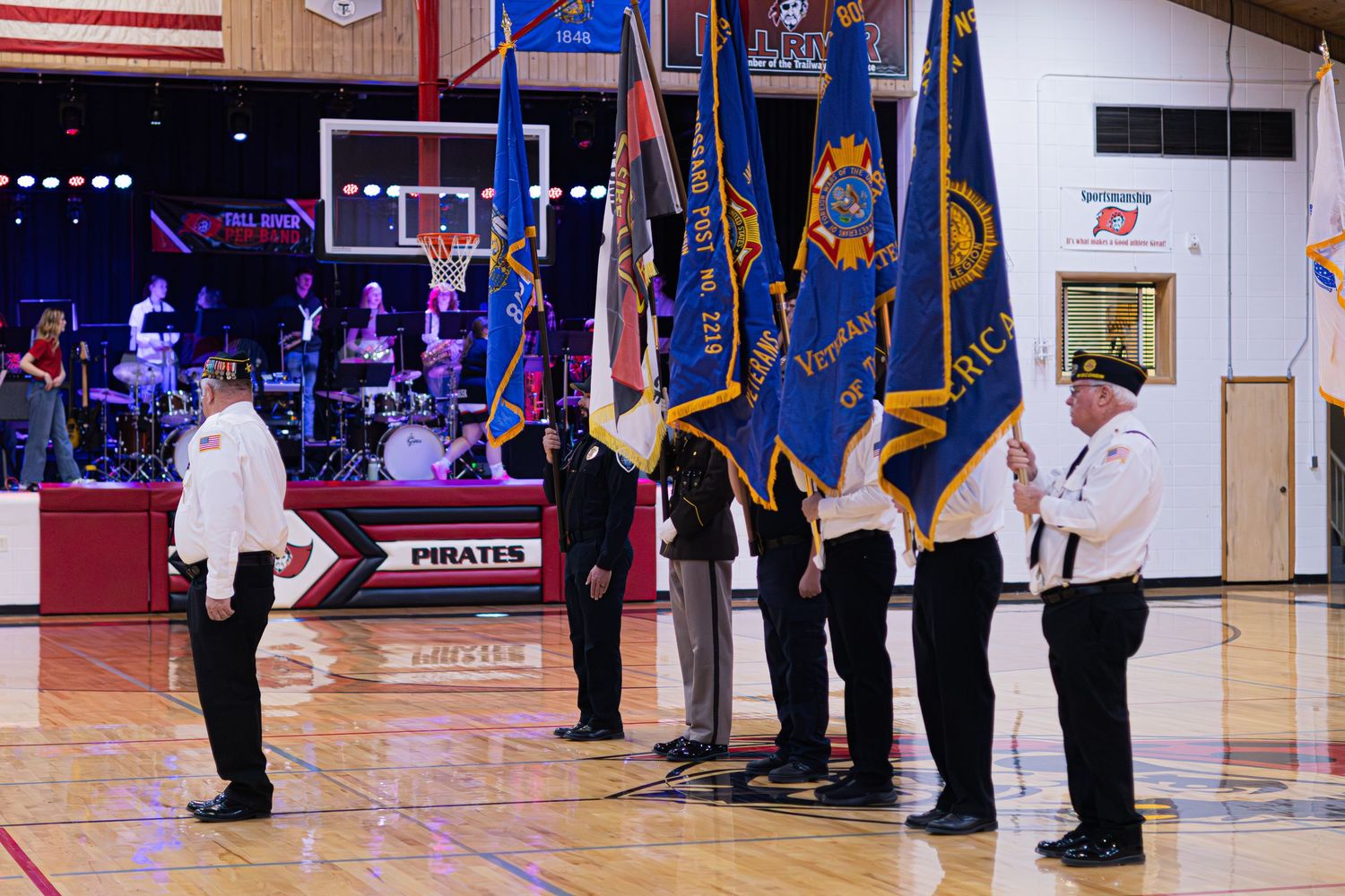 Uniformed service members stand in formation during a military ceremony under an American flag in a gymnasium.