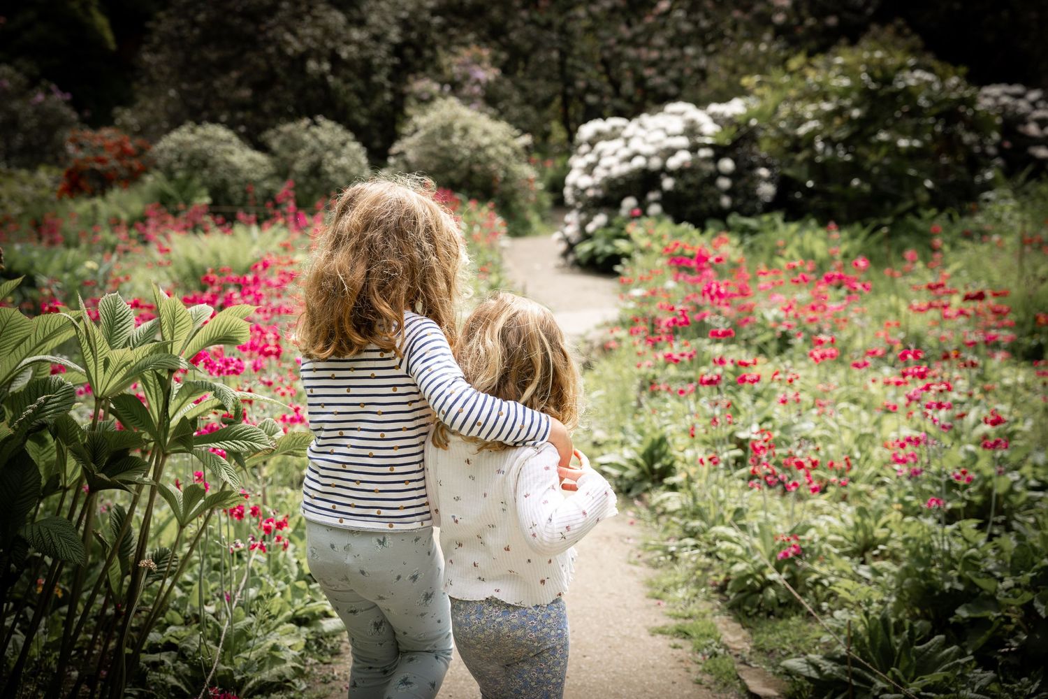 two children with their arms around each other walking down a garden path