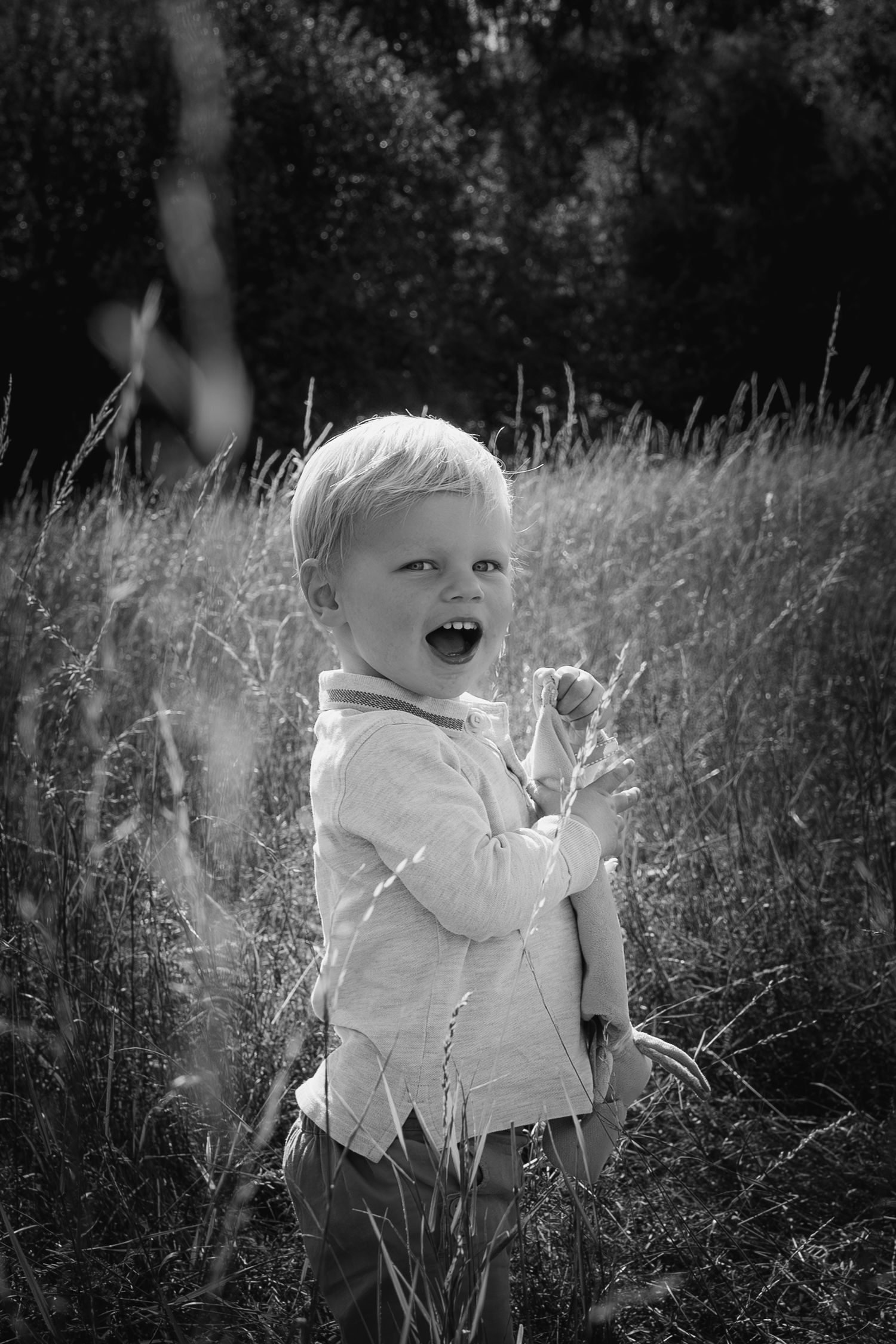 little boy standing in a long grassy meadow, holding a soft toy and laughing