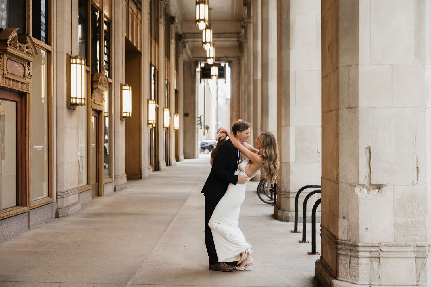Couple sharing a romantic dip kiss under elegant archway columns in a grand city building corridor.