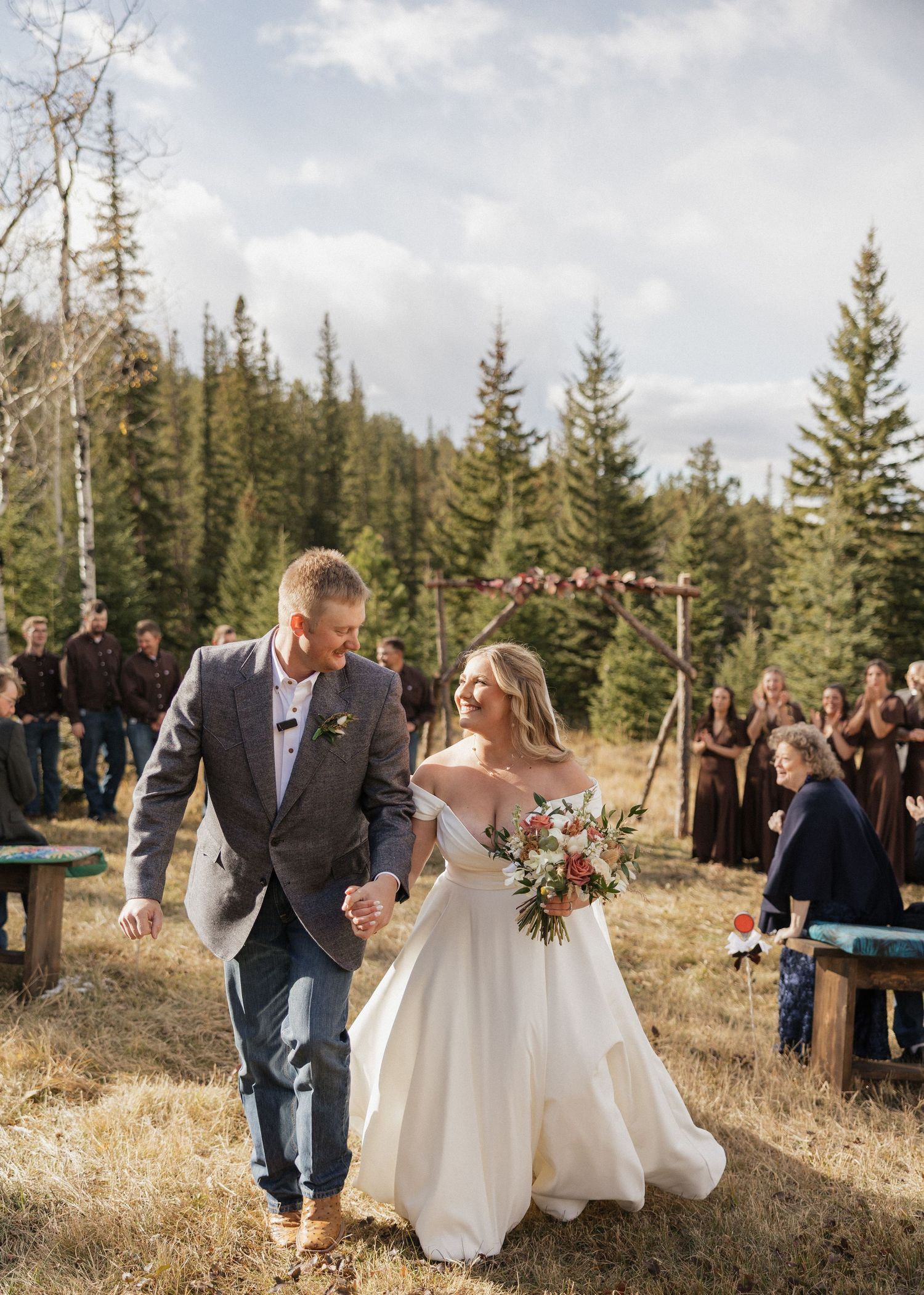 Newlywed couple walks together on a mountain trail surrounded by evergreen trees and wedding guests.