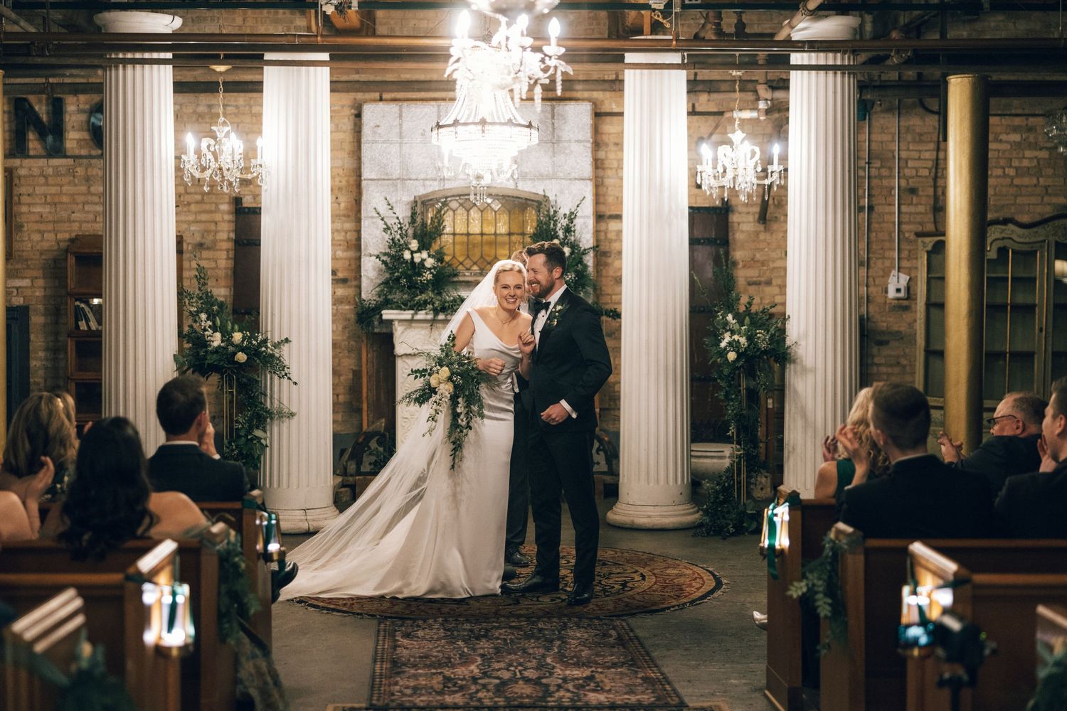 A wedding couple shares their first dance under elegant crystal chandeliers in a grand ballroom with white columns.