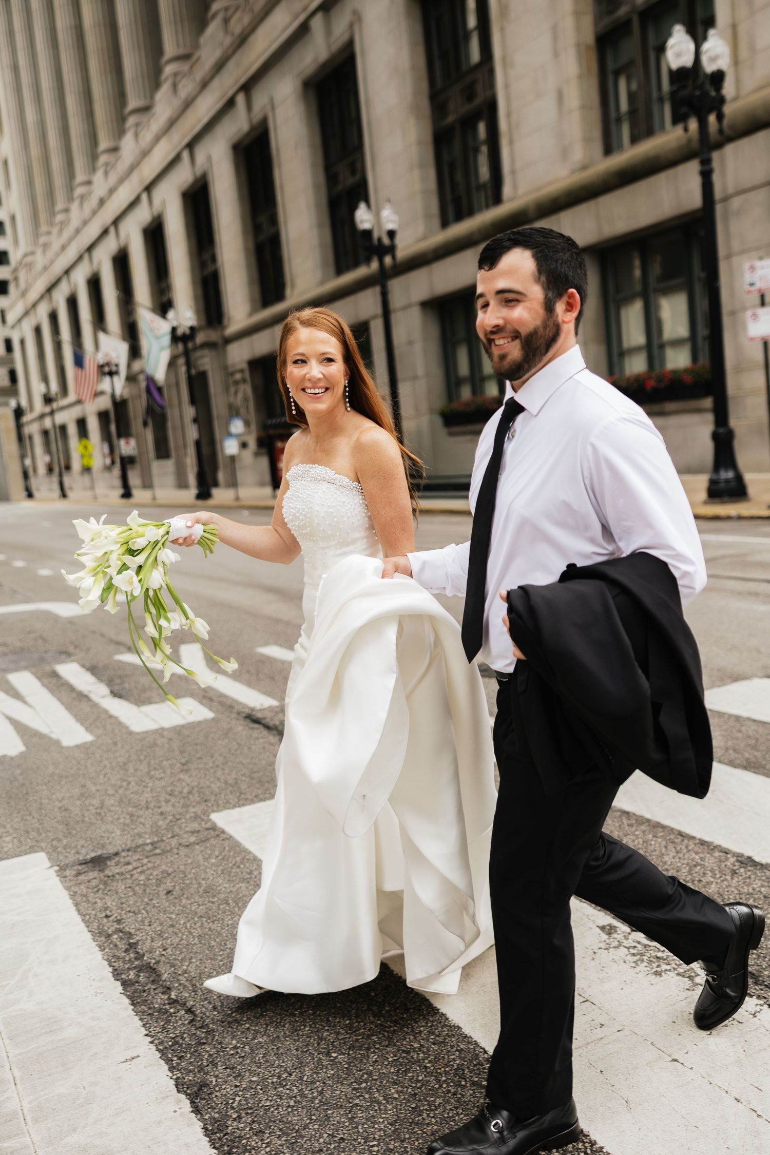 A joyful bride and groom cross a city street, she in a white gown holding flowers, he in a black suit carrying his jacket.