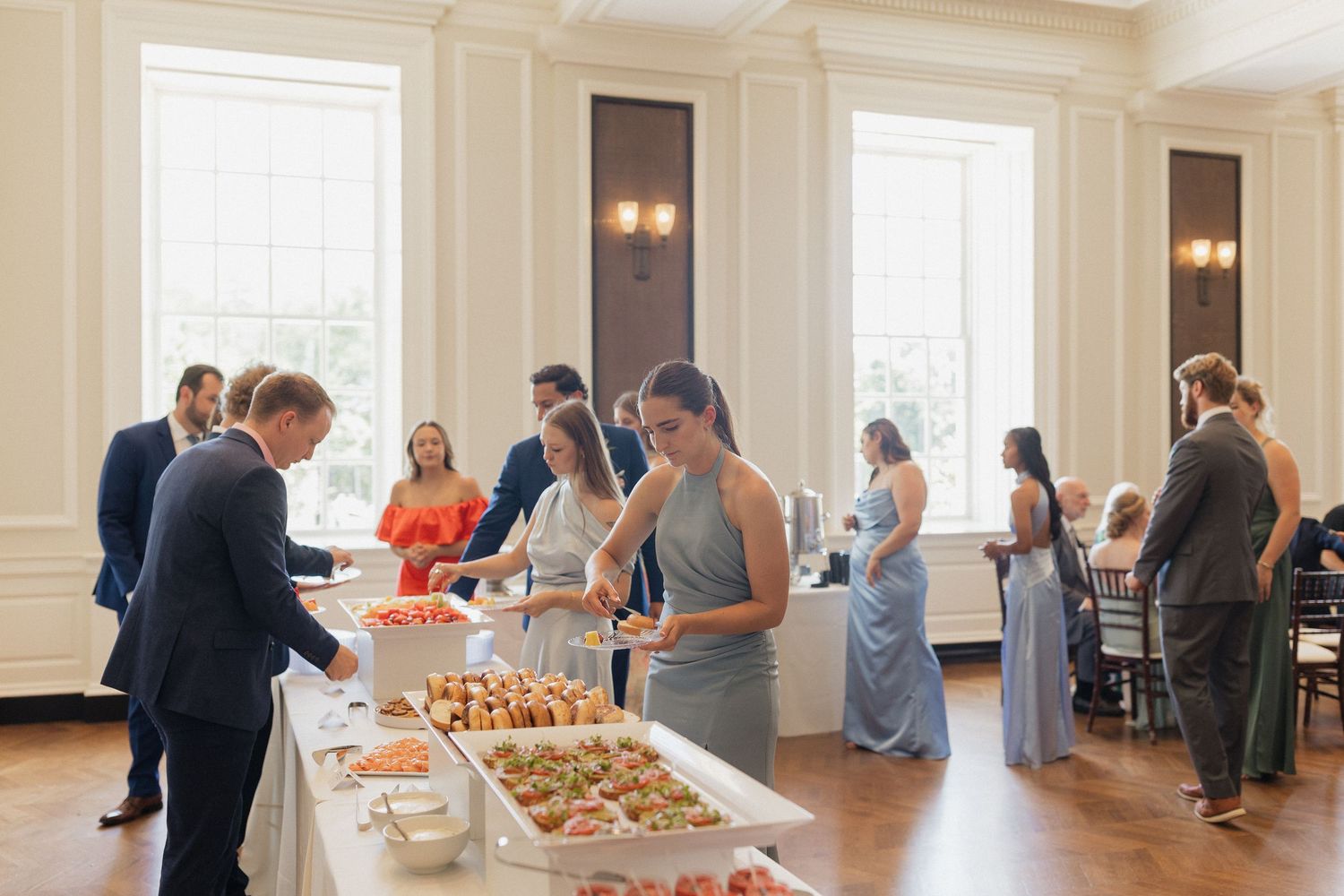Wedding reception guests browse an elegant buffet spread in a bright, white-walled venue.