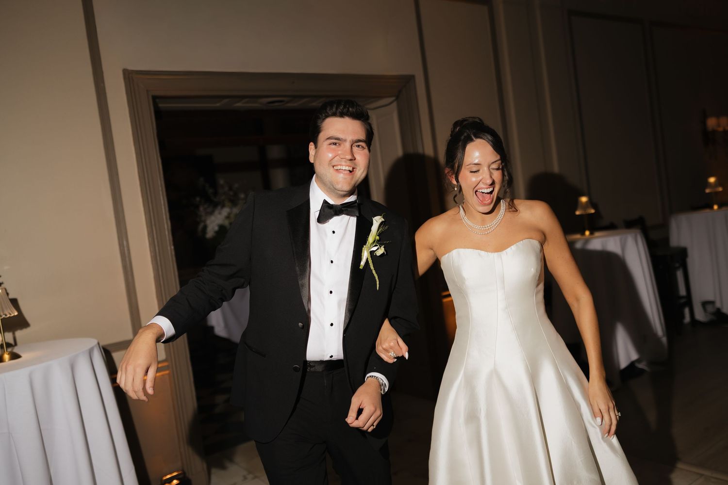 Joyful bride and groom in formal wedding attire holding hands and smiling as they make their grand reception entrance.