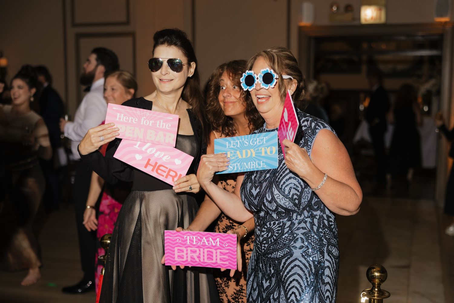 Three women posing with bachelorette party props including Team Bride sign and fun glasses at an indoor event.