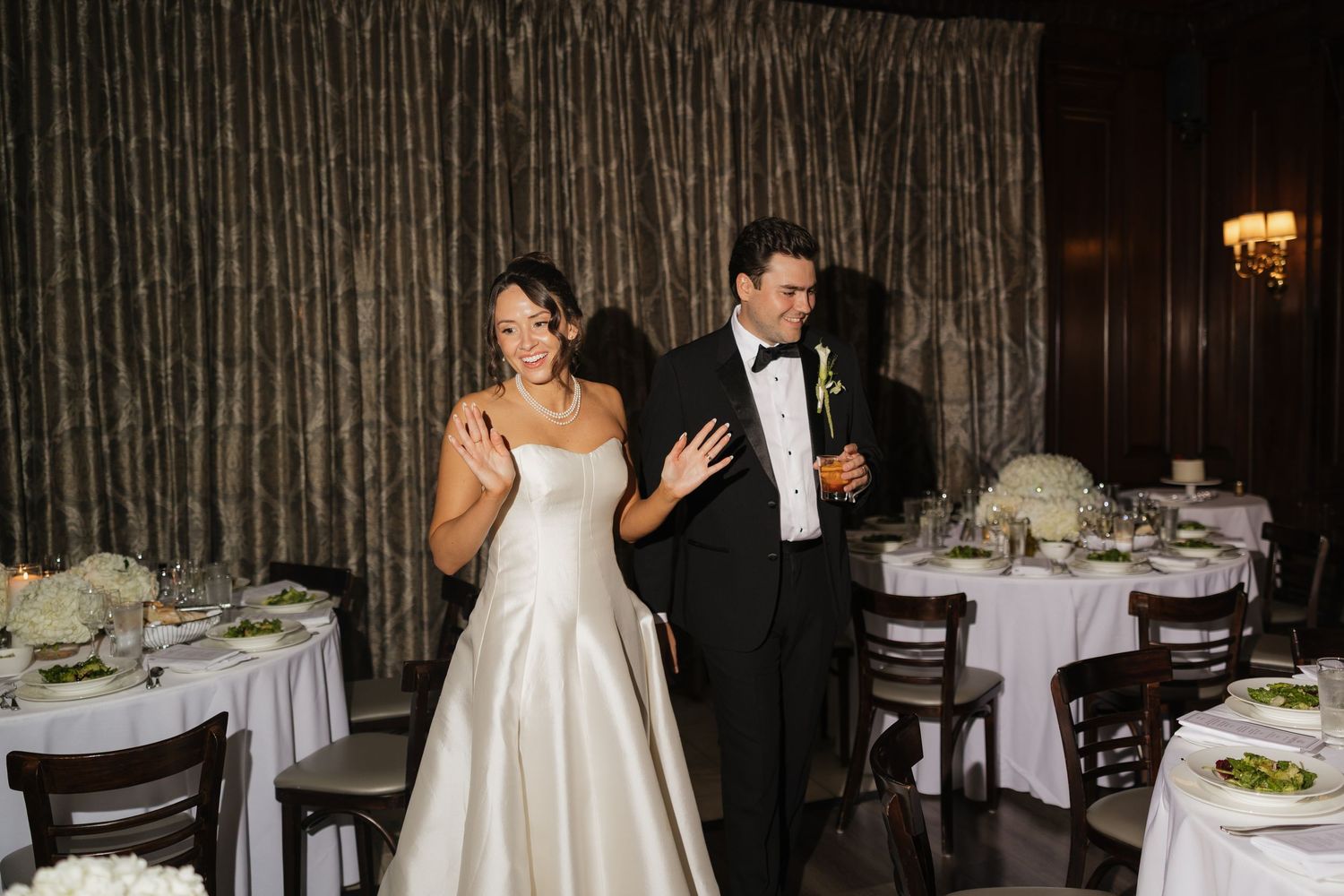 Bride in white satin gown and groom in tuxedo entering elegant wedding reception hall with draped curtains.