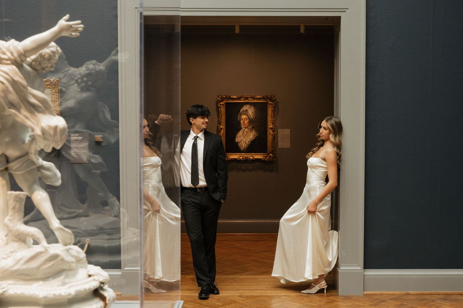 Couple in formal attire posing in a museum doorway with classical sculptures and portrait painting.