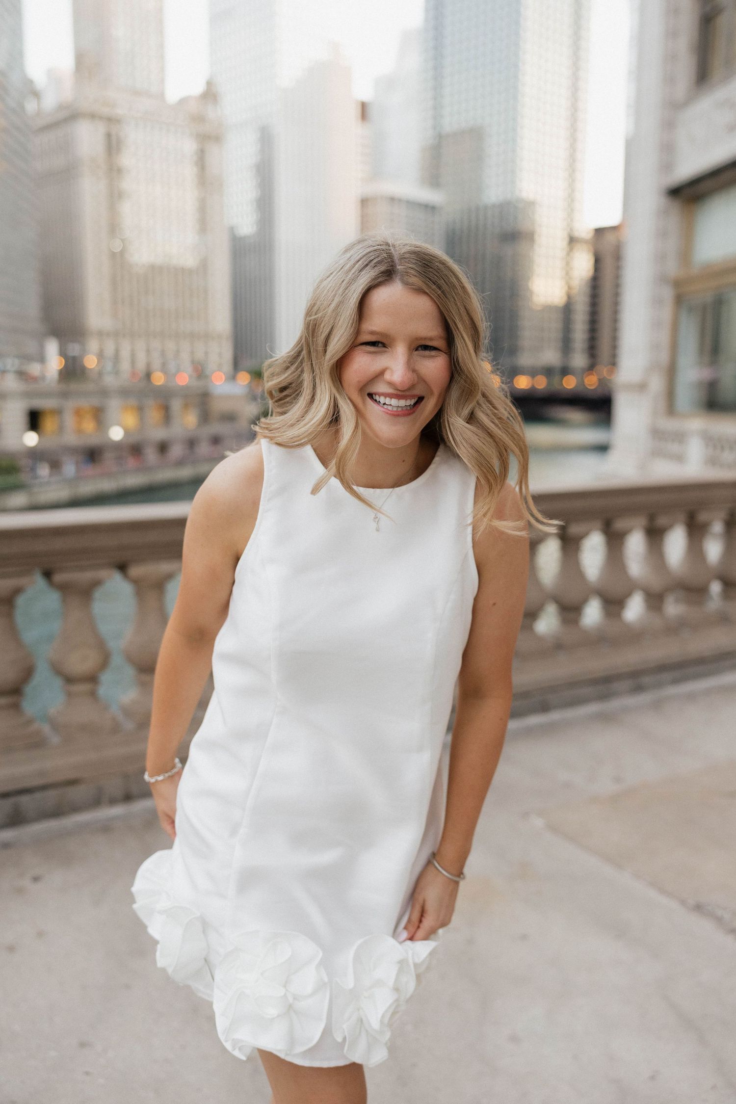 Smiling blonde woman in white sleeveless mini dress on Chicago riverwalk with city skyline behind.