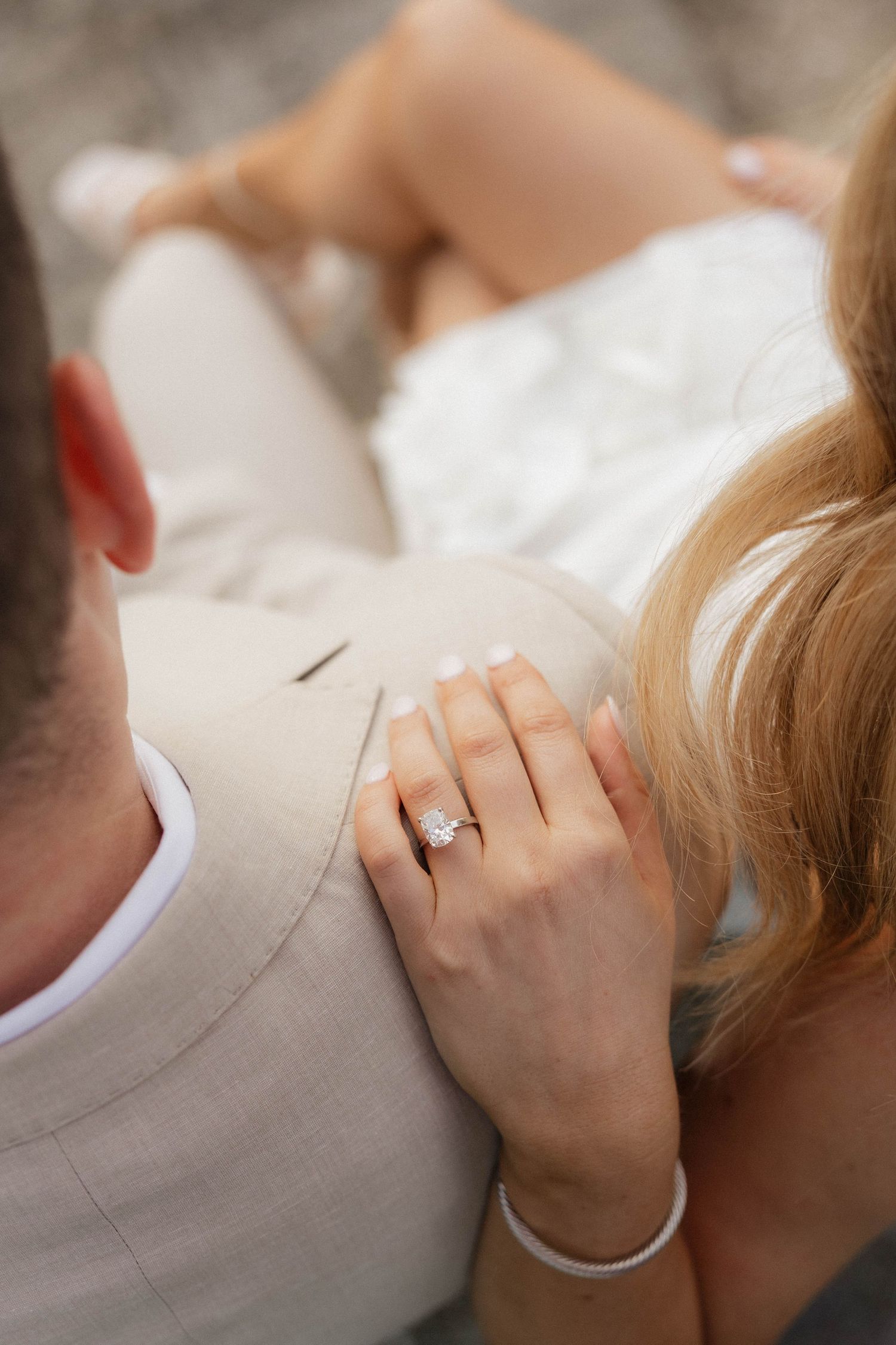Close-up of womans hand with diamond engagement ring resting on mans beige suit jacket.