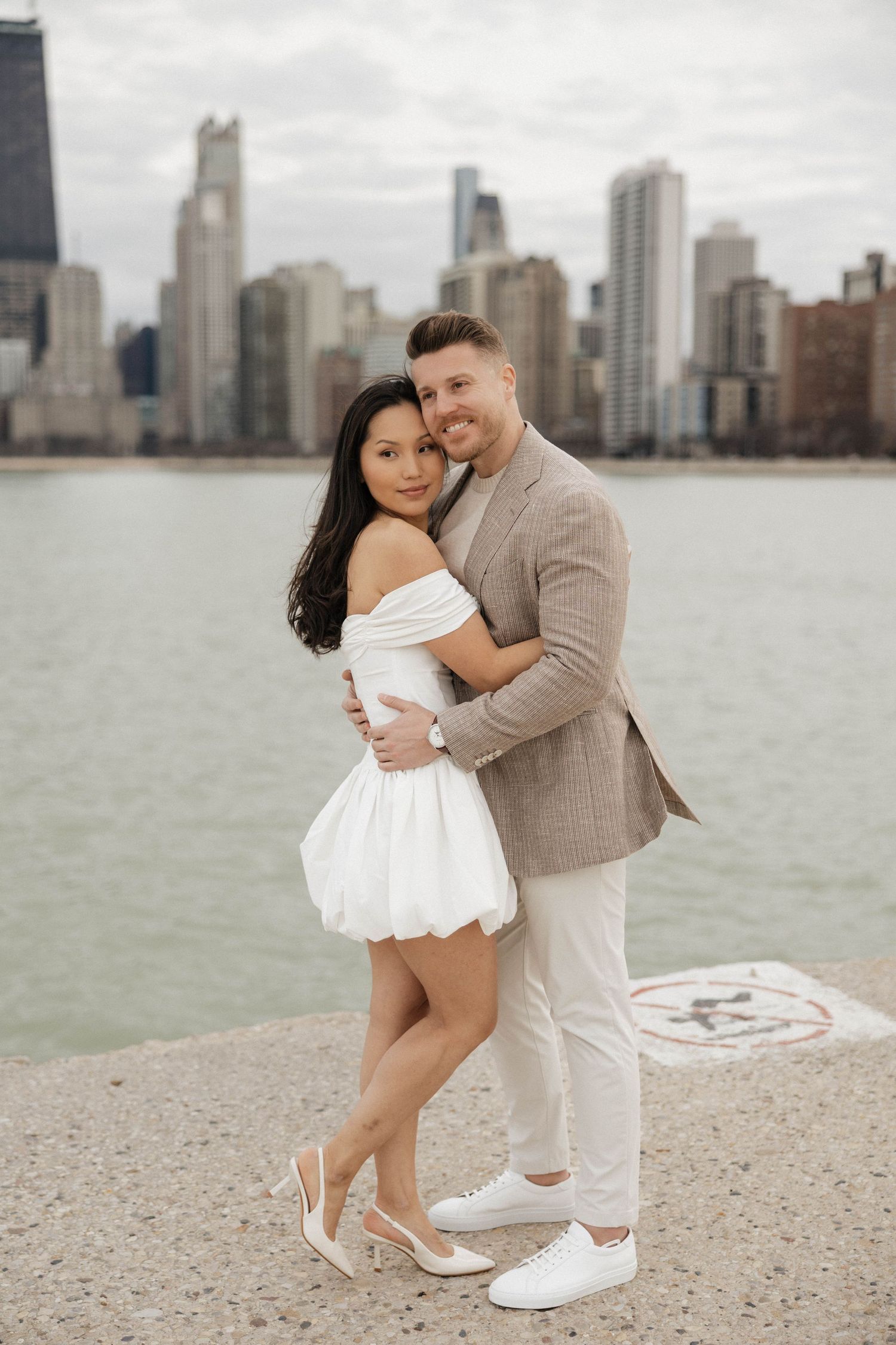 Couple embracing on Chicago waterfront, woman in white off-shoulder dress, man in beige blazer, city skyline backdrop.