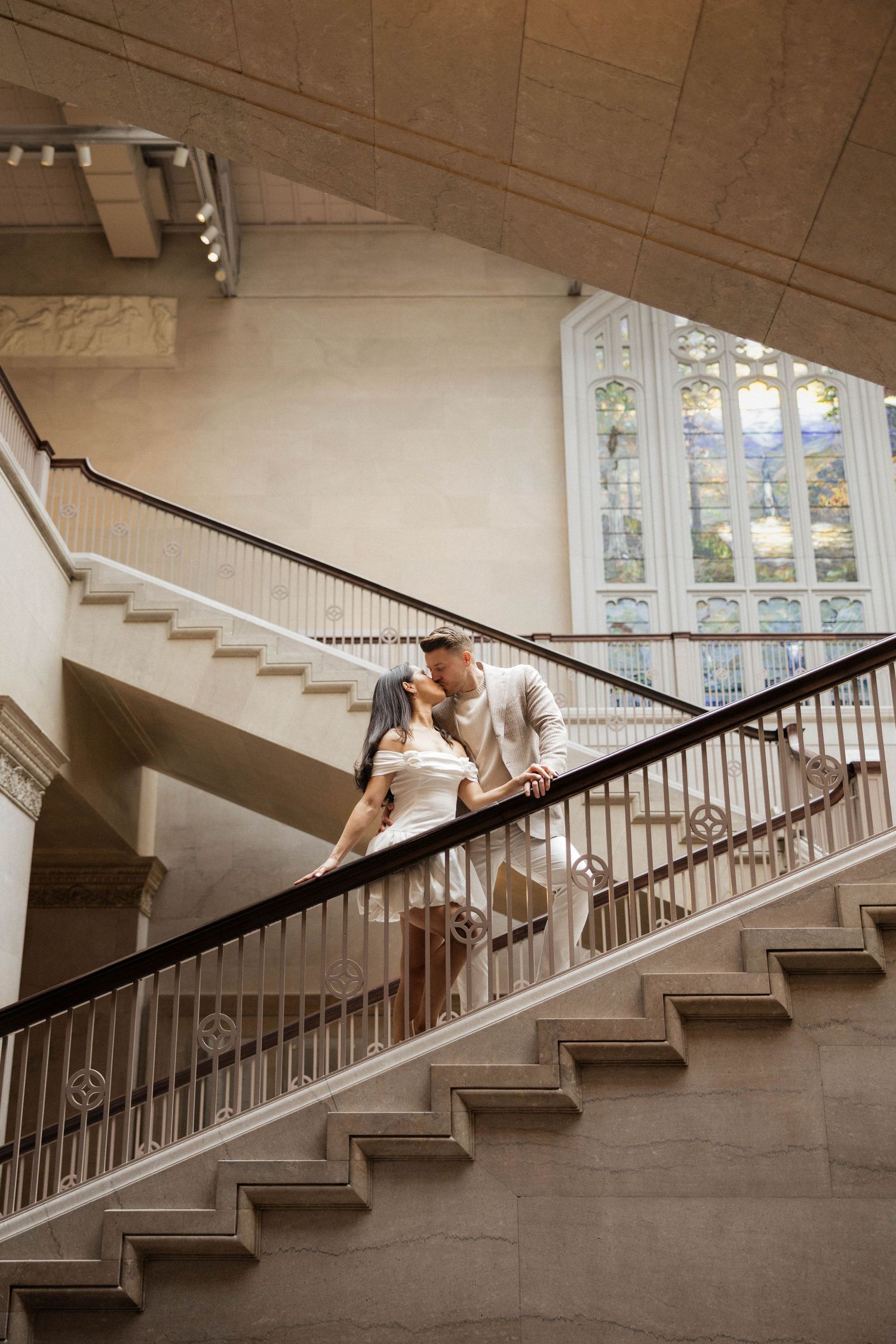 Romantic couple kissing on grand staircase inside elegant museum with ornate windows.